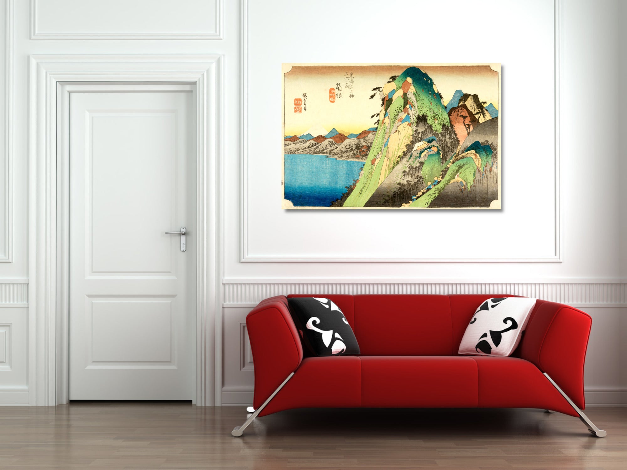 A framed Japanese ukiyo-e style art print depicting a green mountain landscape with blue water, displayed on a white wall above a modern red sofa with black and white patterned pillows.