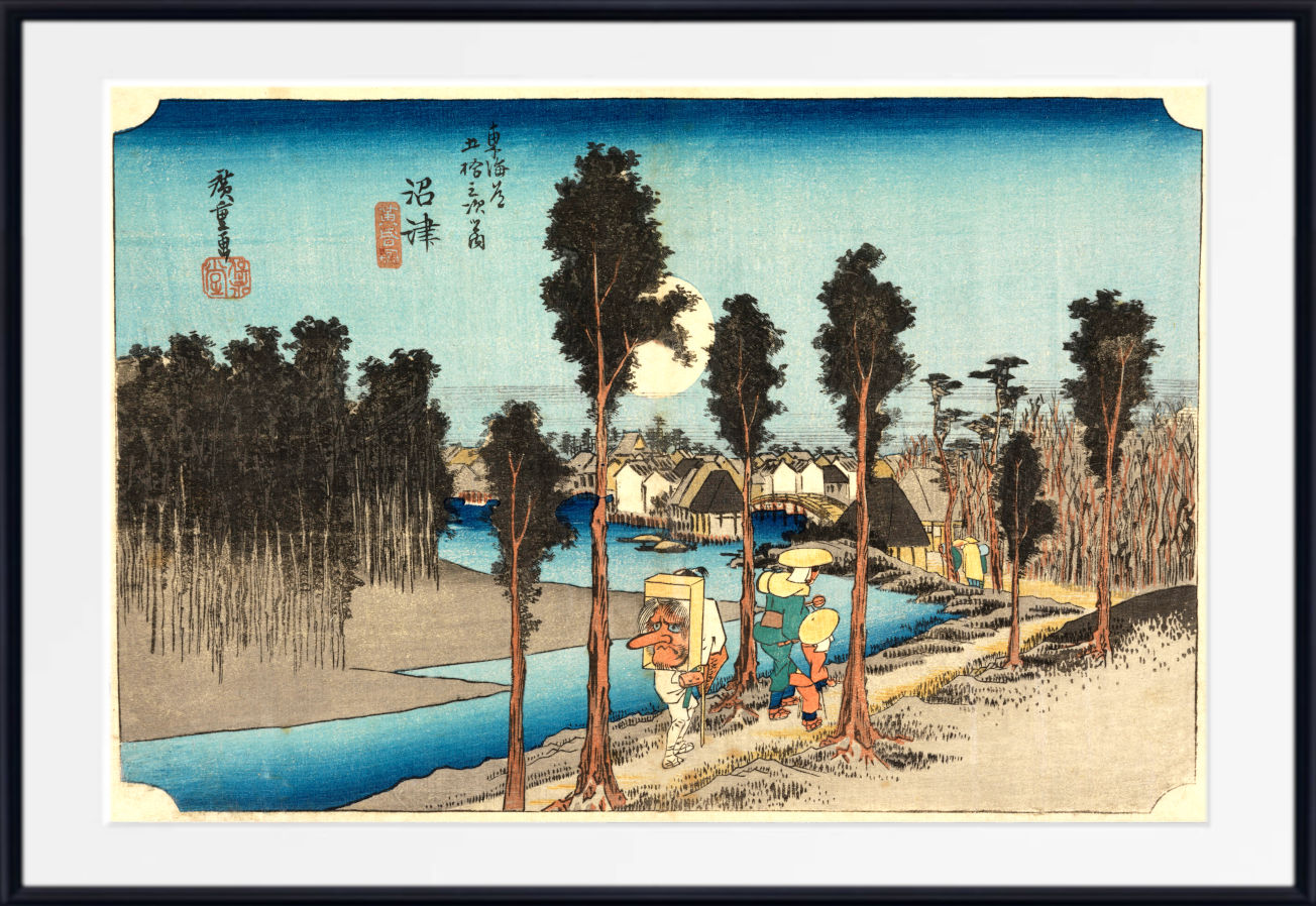 A framed Japanese ukiyo-e woodblock print showing a river scene with tall trees, small boats, houses, and a group of people walking along a path under a pale sky. The print is mounted with a white mat and a black frame.