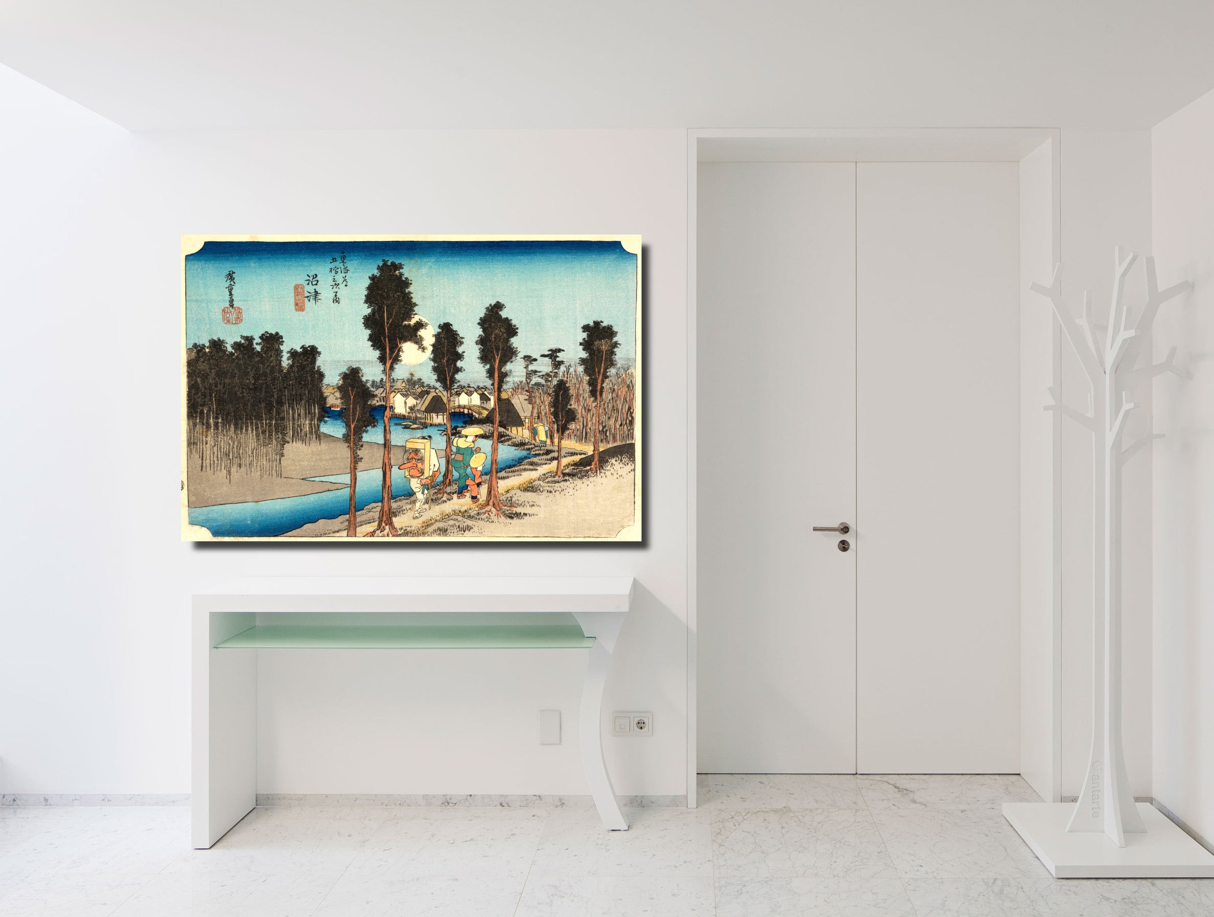 A framed Japanese-style landscape art print hangs on a white wall above a white console table in a bright, modern room. The artwork depicts a river scene with trees, boats, and buildings in blue, beige, and brown tones.