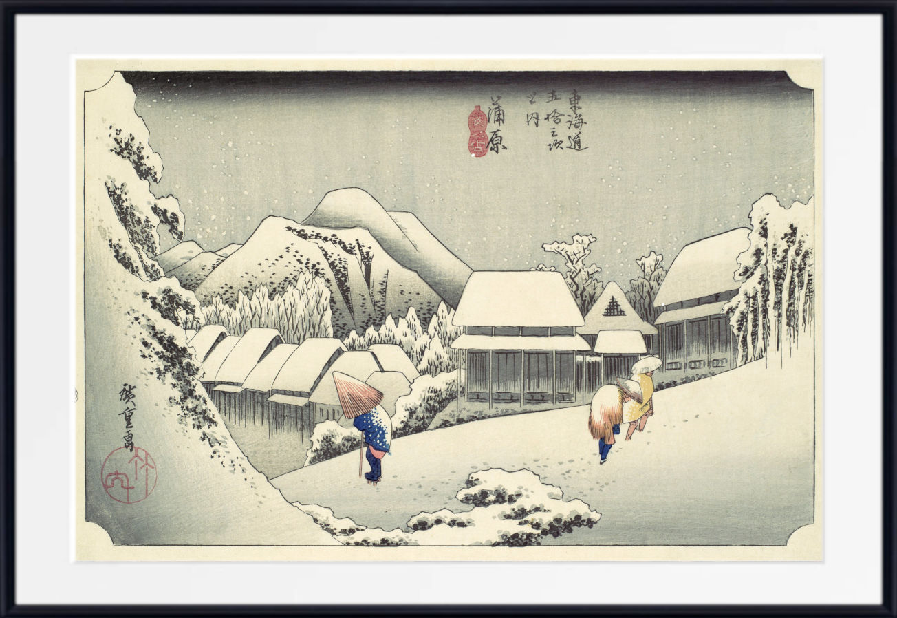 A framed Japanese-style winter landscape print showing snow-covered rooftops, mountains, and two figures walking outdoors, with a white mat and black frame around the artwork.