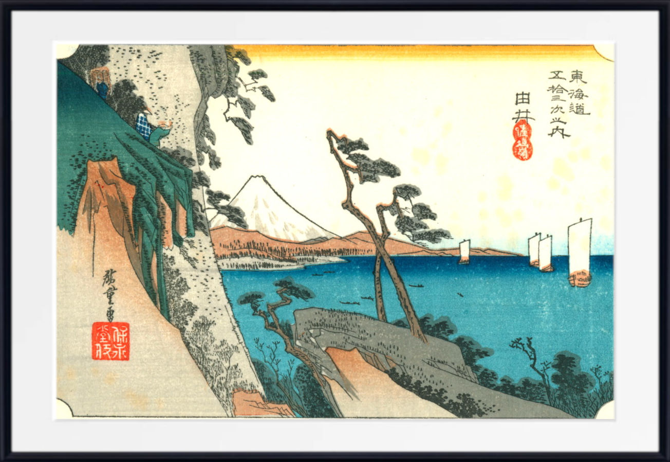 A framed Japanese ukiyo-e woodblock print depicting a coastal landscape with Mount Fuji, blue water, trees, and red seal marks, shown against a white mat and black frame.