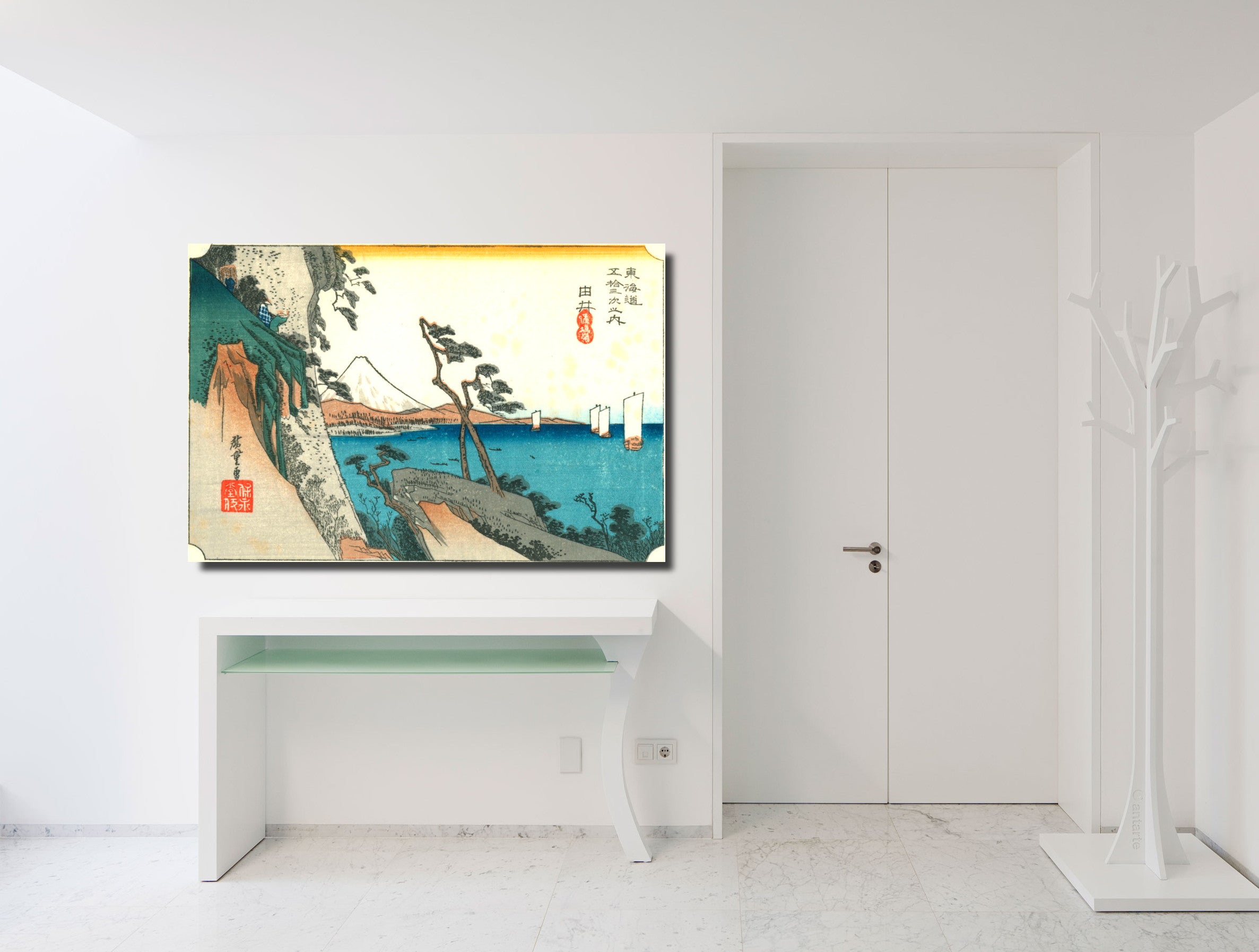 A framed Japanese ukiyo-e style landscape art print hangs on a white wall above a white console table in a bright, modern room. The print depicts a scenic coastal landscape with mountains, trees, and blue water, with a prominent yellow band at the top.