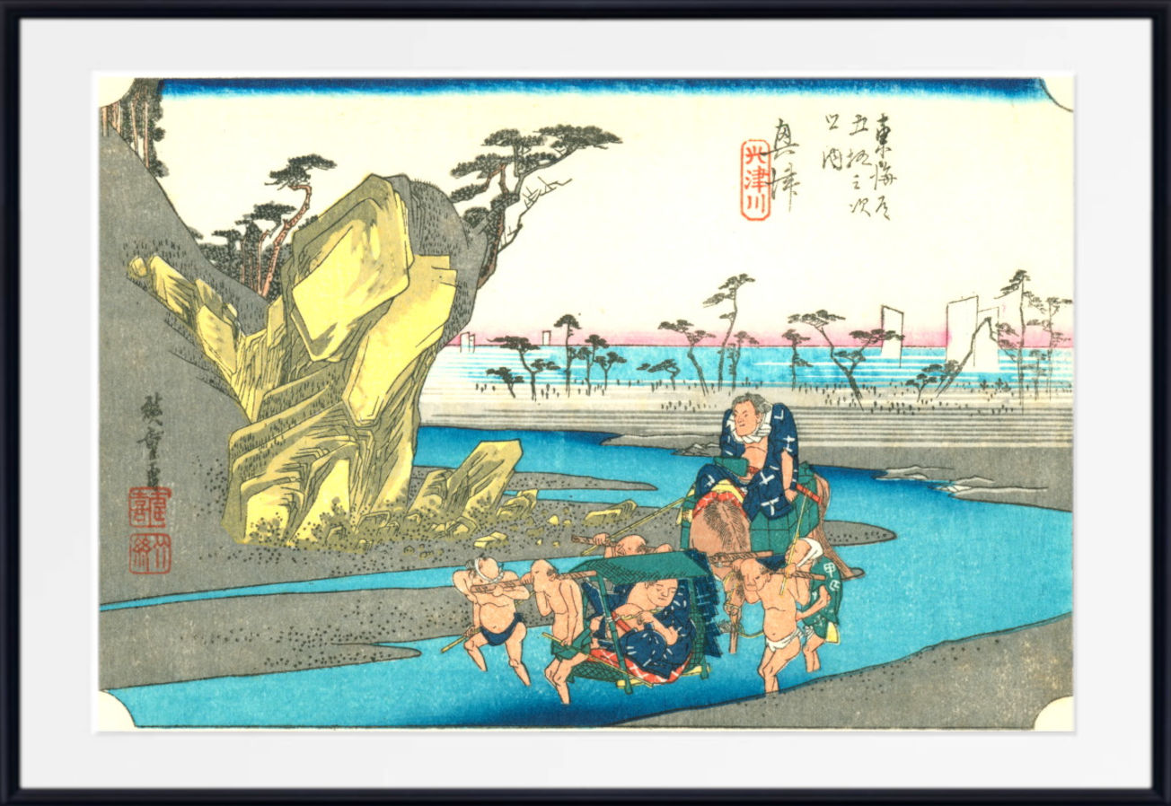A framed Japanese ukiyo-e woodblock print depicting a coastal scene with figures in a river, a large yellow rock formation, trees, and distant buildings, shown against a white mat and black frame.