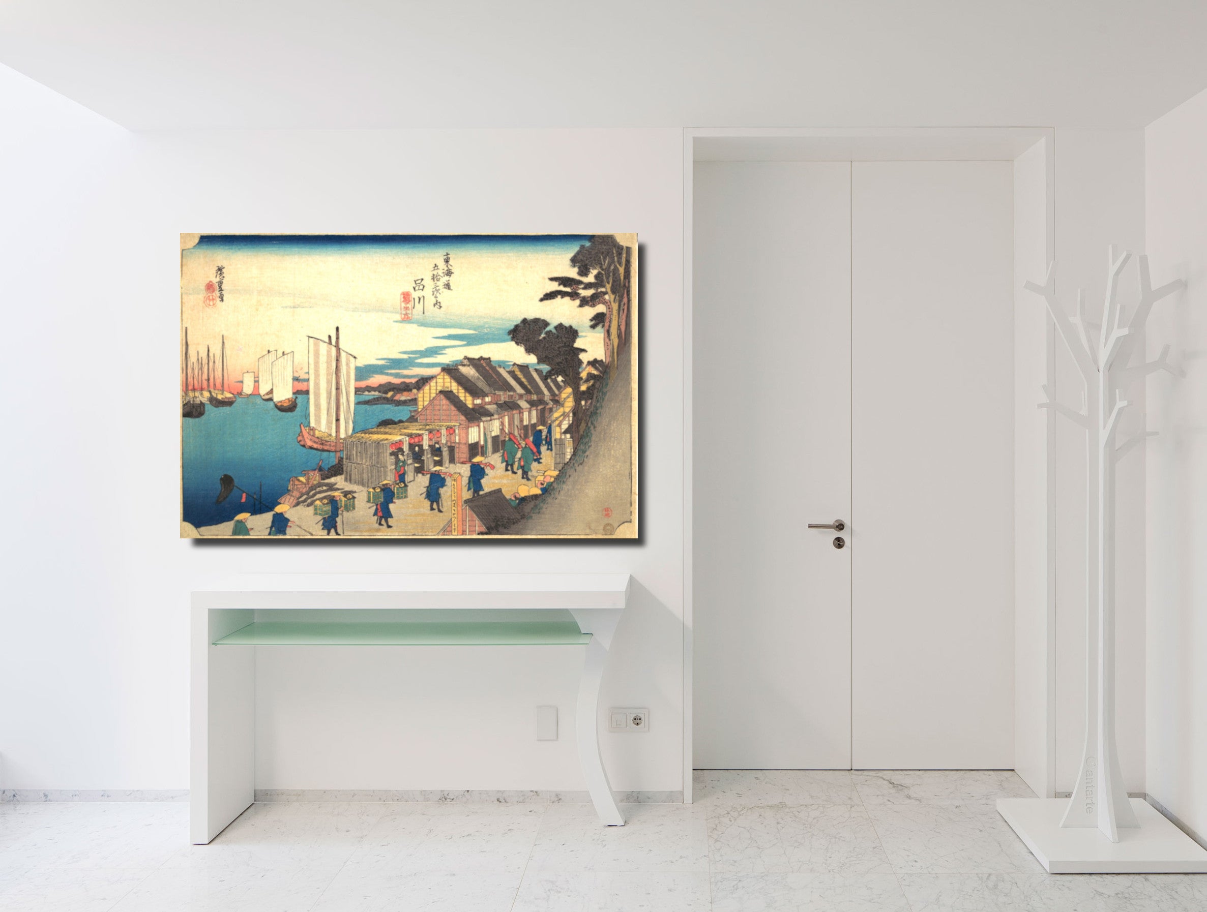 A framed Japanese ukiyo-e woodblock print depicting a harbor scene with boats, figures, and buildings, displayed on a white wall above a white console table in a bright minimalist interior.