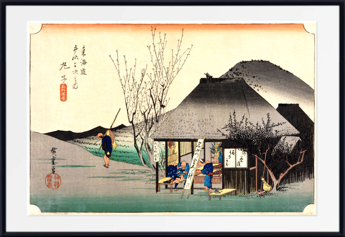 A framed Japanese ukiyo-e woodblock print showing a rural scene with a thatched-roof house, figures near a doorway, bare trees, and a mountain in the background. The print is centered within a white mat and a black frame.