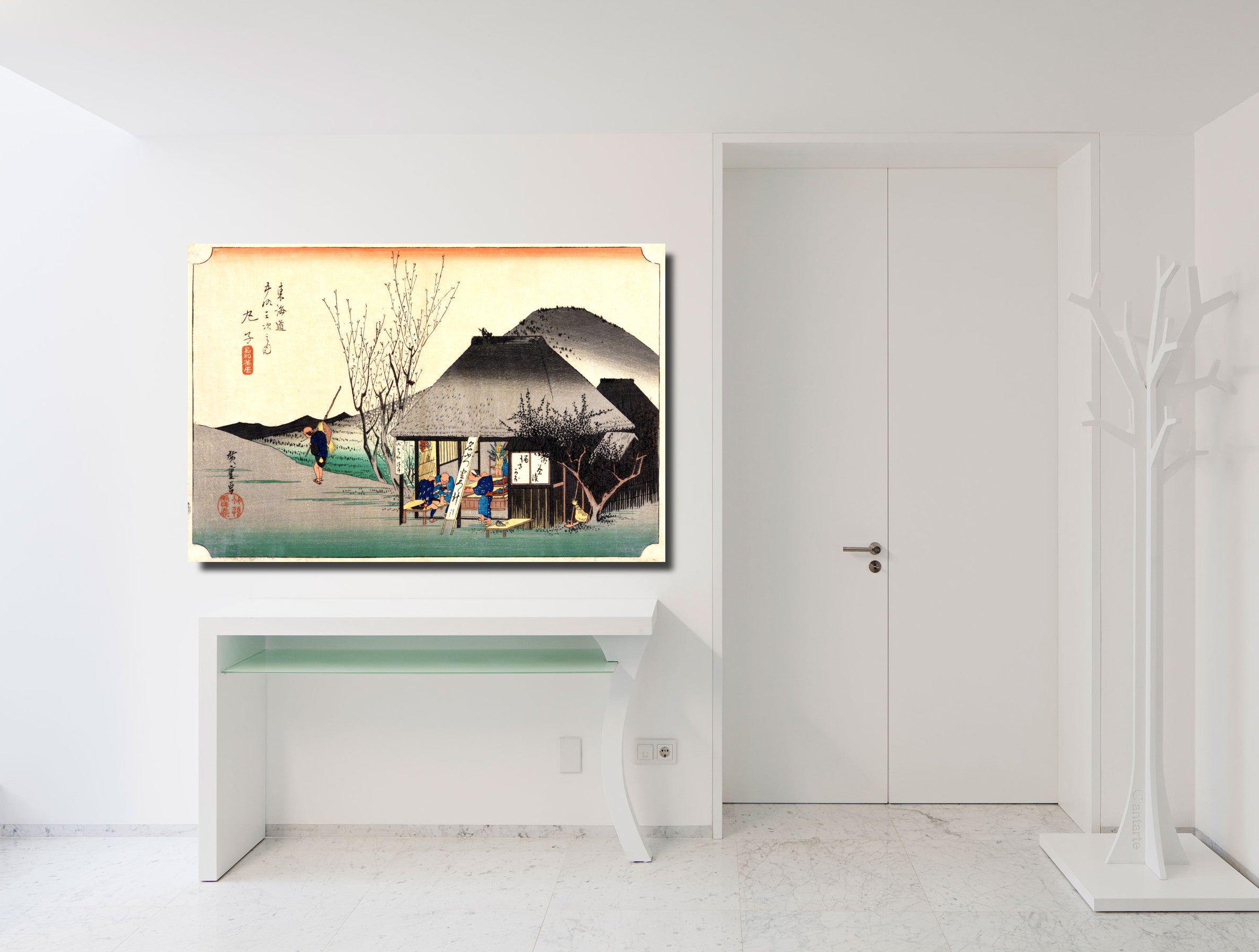 A framed Japanese-style art print depicting a traditional village scene with a thatched-roof house, tree branches, and mountains, displayed on a white wall above a white console table in a bright modern room.