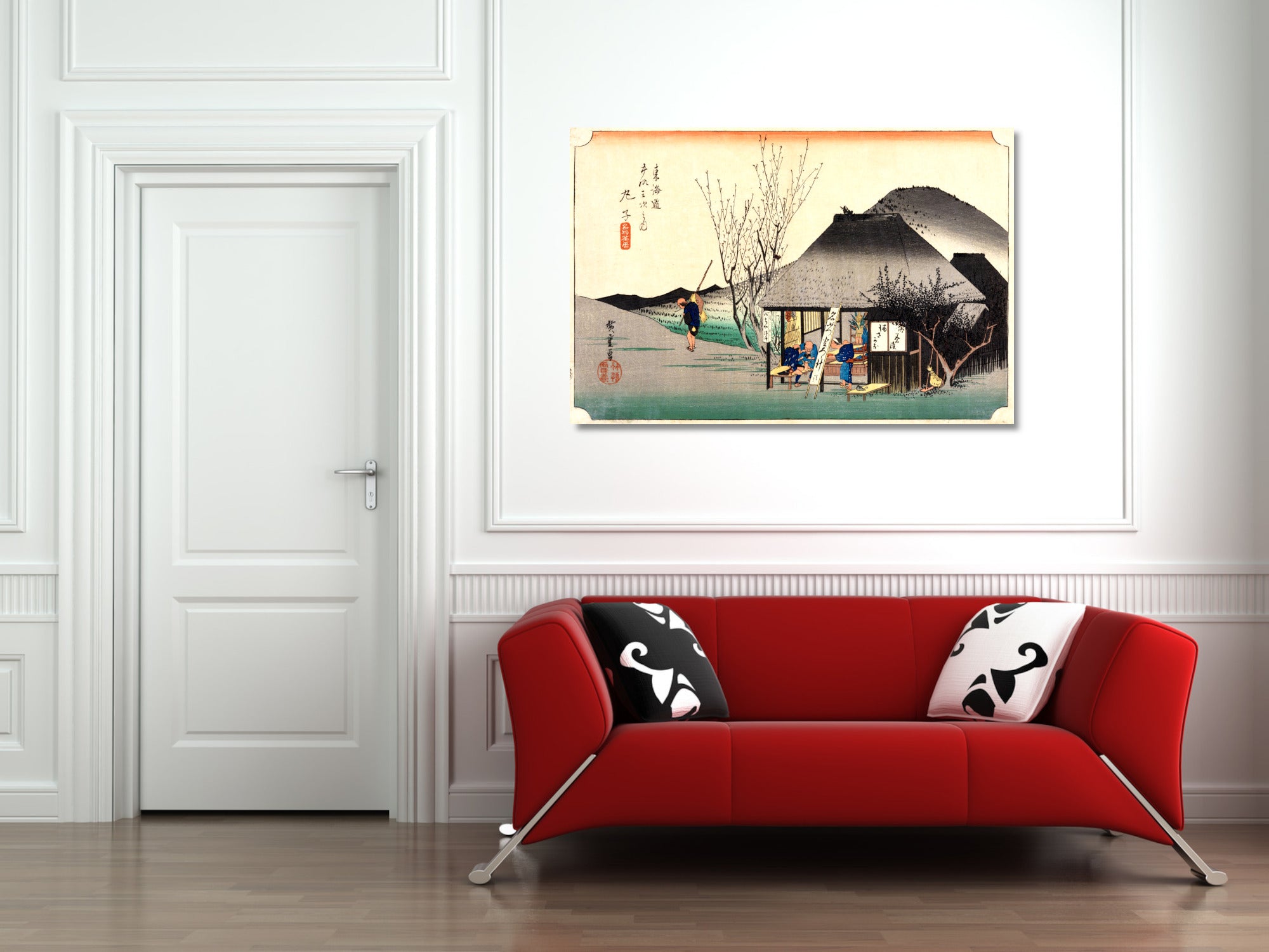 A framed Japanese-style art print depicting a traditional village scene with a thatched house and mountain, displayed on a white wall above a modern red sofa with black-and-white patterned pillows.
