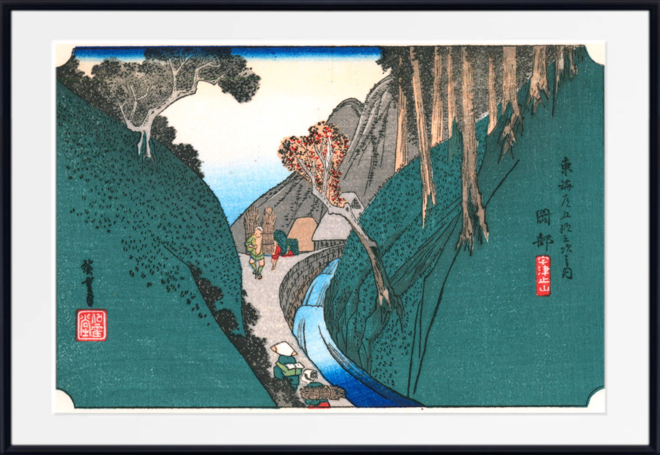 A framed Japanese ukiyo-e woodblock print depicting a mountain valley with a winding blue river, trees, and small figures, shown with a white mat and black frame.