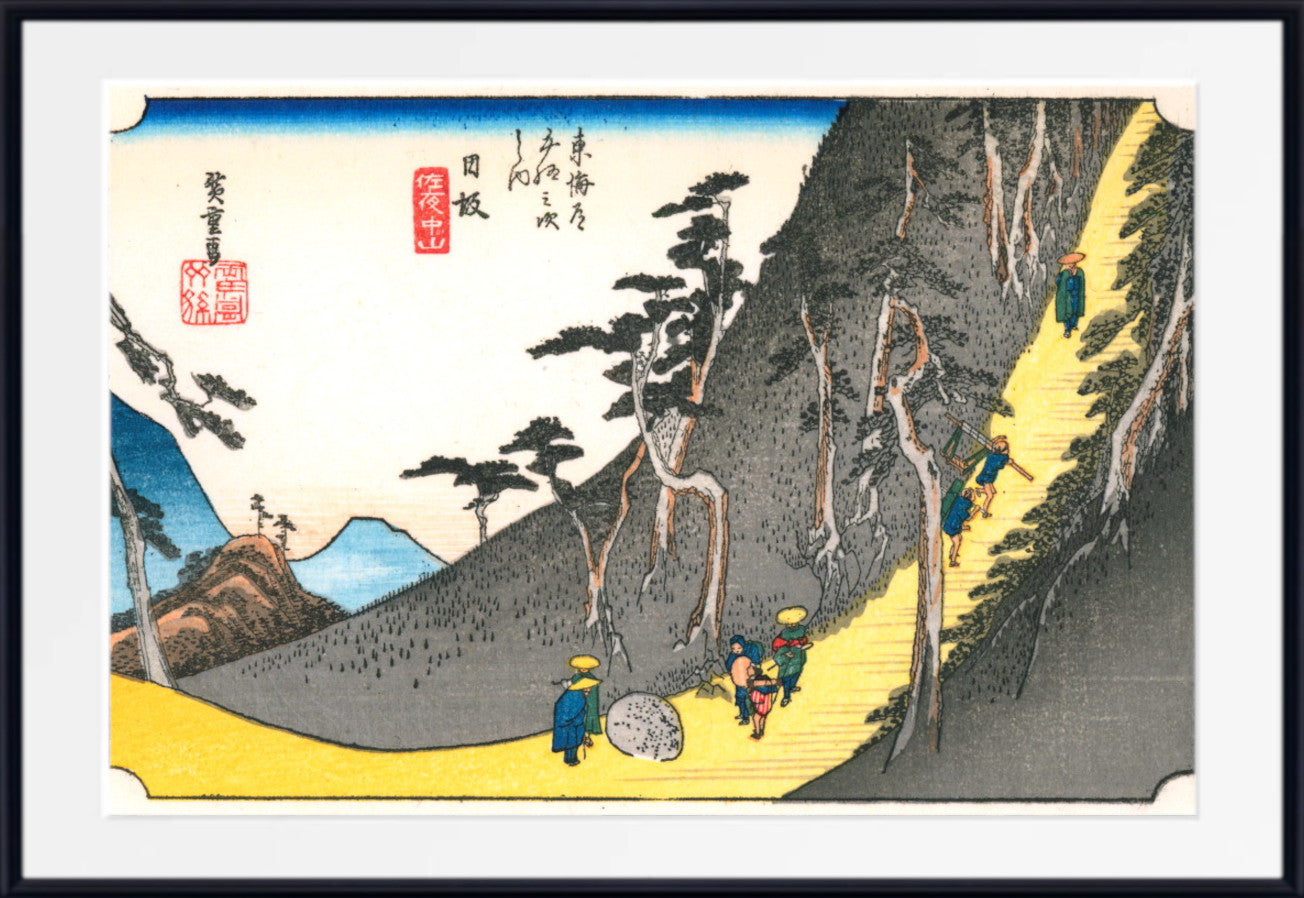 A framed Japanese-style art print depicting a mountain path with small figures walking along a yellow road, trees, blue water, and red seal marks, shown against a white mat and black frame.