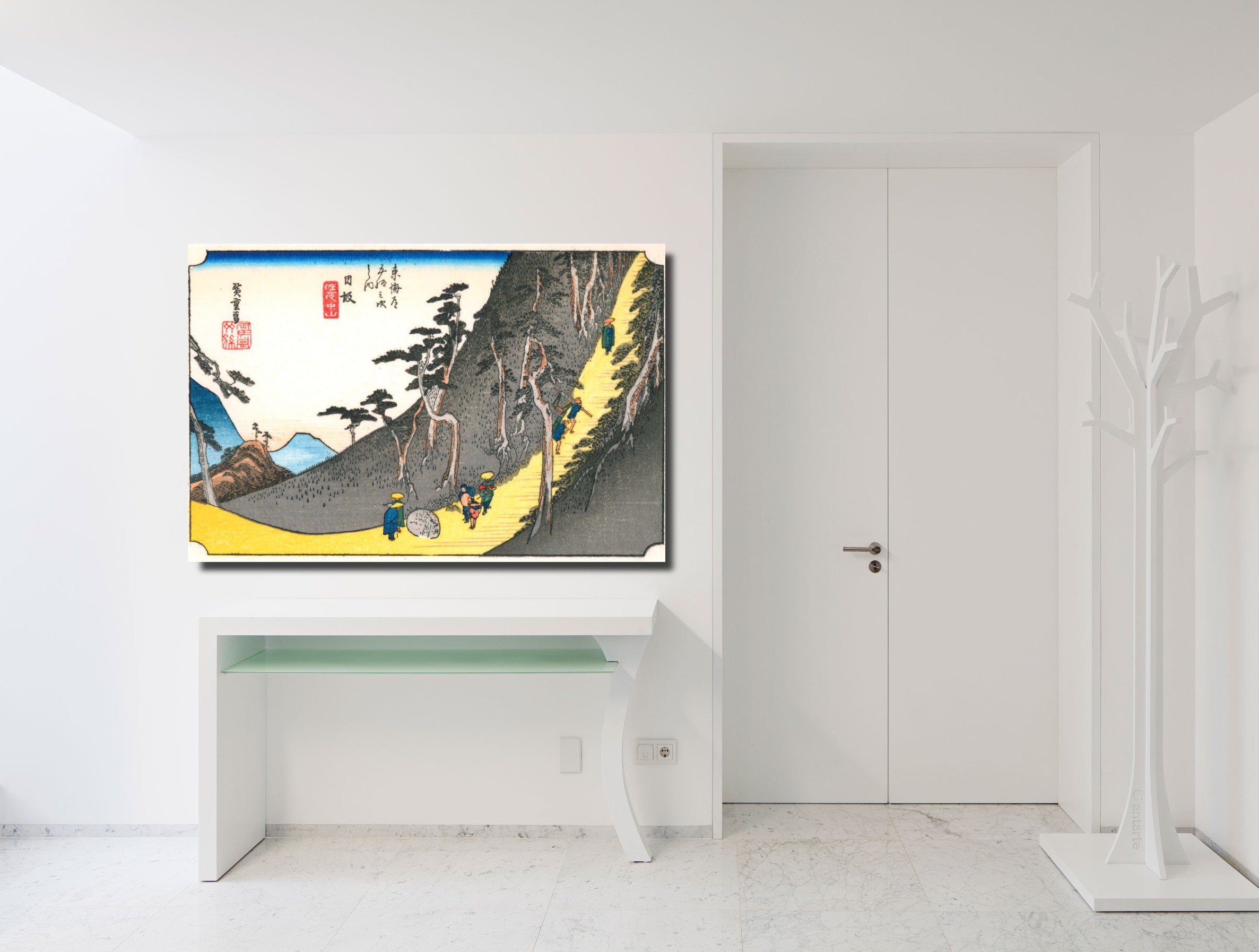 A framed art print of a Japanese ukiyo-e woodblock scene, depicting a mountain landscape with figures, displayed on a white wall above a white console table in a minimalist room.