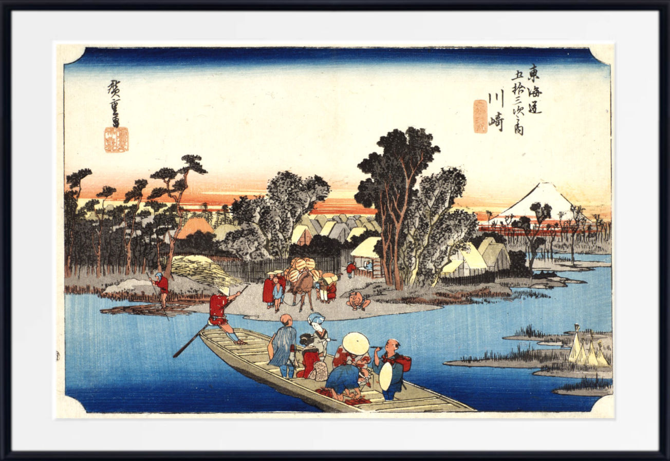 A framed Japanese ukiyo-e woodblock print showing a river scene with a boat, trees, buildings, and a mountain at sunset, presented with a white mat and black frame.