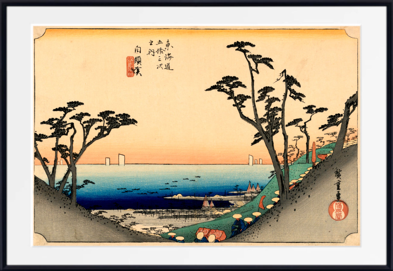 A framed Japanese ukiyo-e woodblock print depicting a coastal landscape with pine trees, a blue sea, and a pale peach sky, shown with a white mat and black frame.