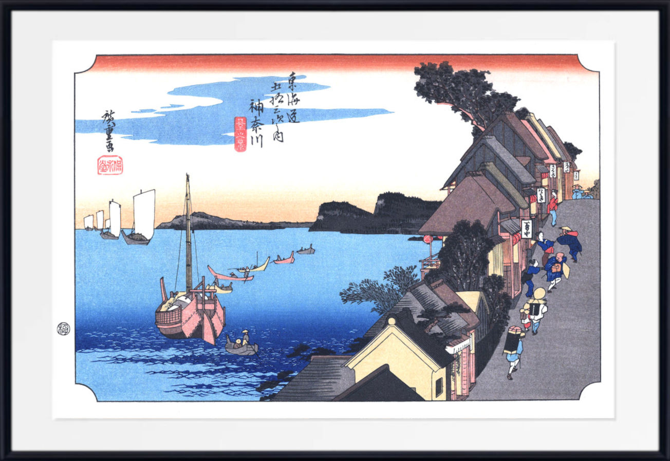 A framed Japanese ukiyo-e woodblock print depicting a coastal harbor scene with boats on blue water, buildings along the shore, and a pale sky with red-orange clouds. The print is shown with a white mat and a black frame.