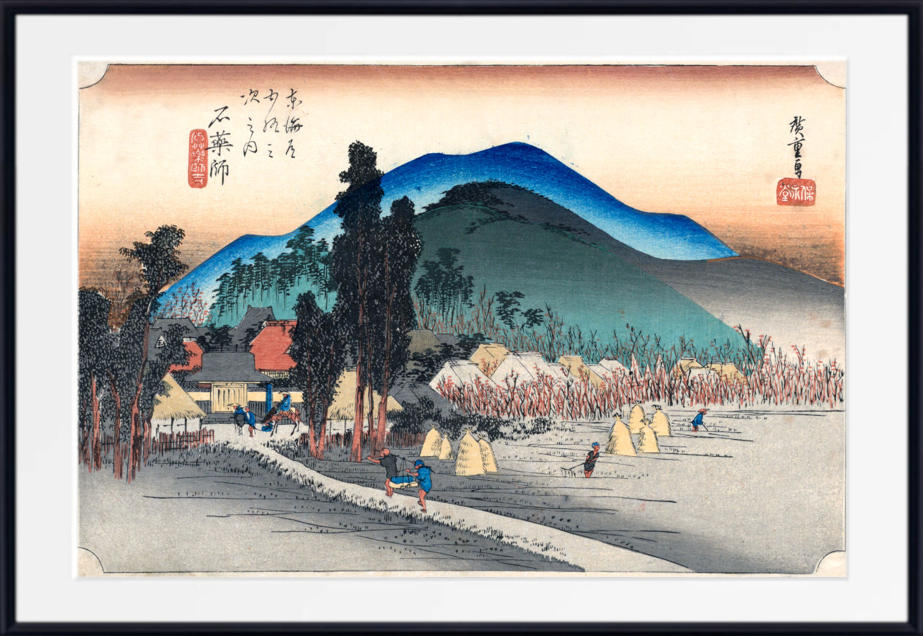 A framed Japanese ukiyo-e woodblock print showing a rural landscape with mountains, trees, houses, and figures in the foreground, presented with a white mat and black frame.