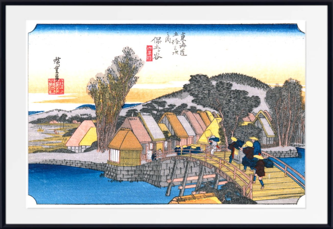 A framed Japanese ukiyo-e woodblock print showing a waterfront village with thatched-roof houses, a wooden bridge, trees, and figures walking under a blue sky. The print is mounted with a white mat and a black frame.