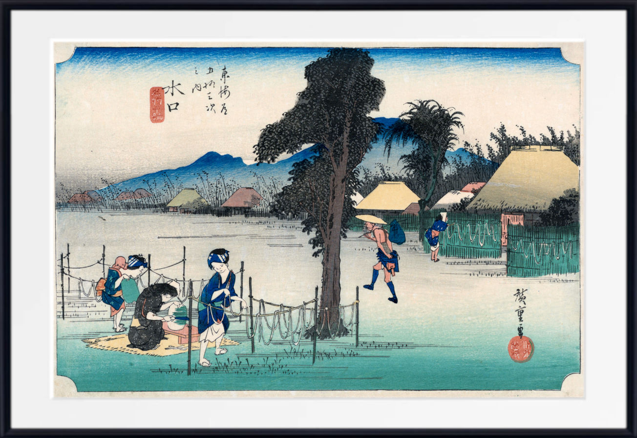A framed Japanese ukiyo-e woodblock print depicting figures by a body of water with trees, thatched-roof houses, and distant mountains. The artwork is shown with a white mat and a black frame.