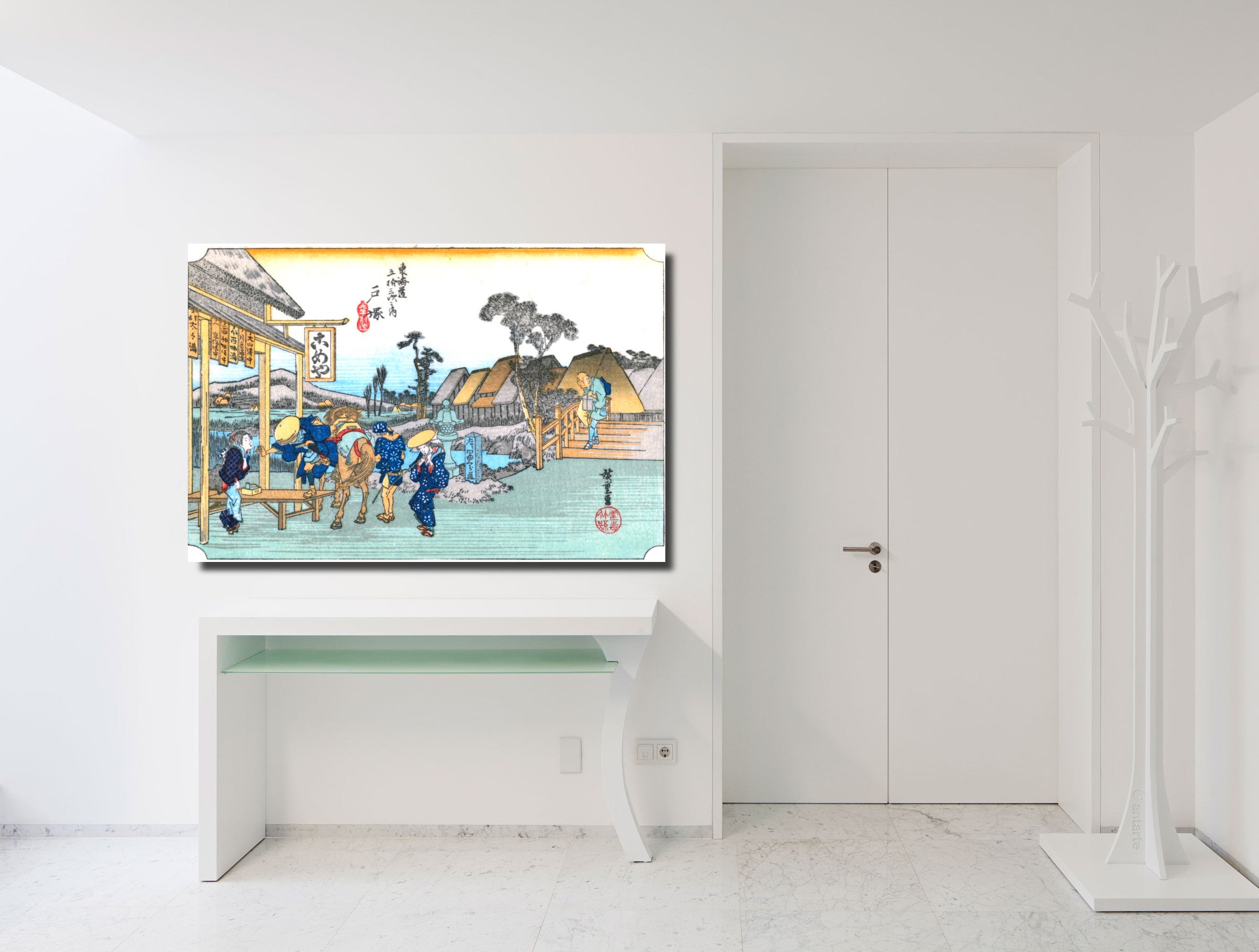 A framed Japanese ukiyo-e woodblock print featuring a street scene with figures, boats, and buildings, displayed on a white console table in a bright, modern white room.