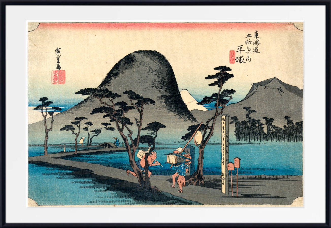 A framed Japanese-style landscape print showing a mountain, water, trees, and two small figures near a post, with red seals and black calligraphy details. The artwork is centered within a white mat and a black frame.