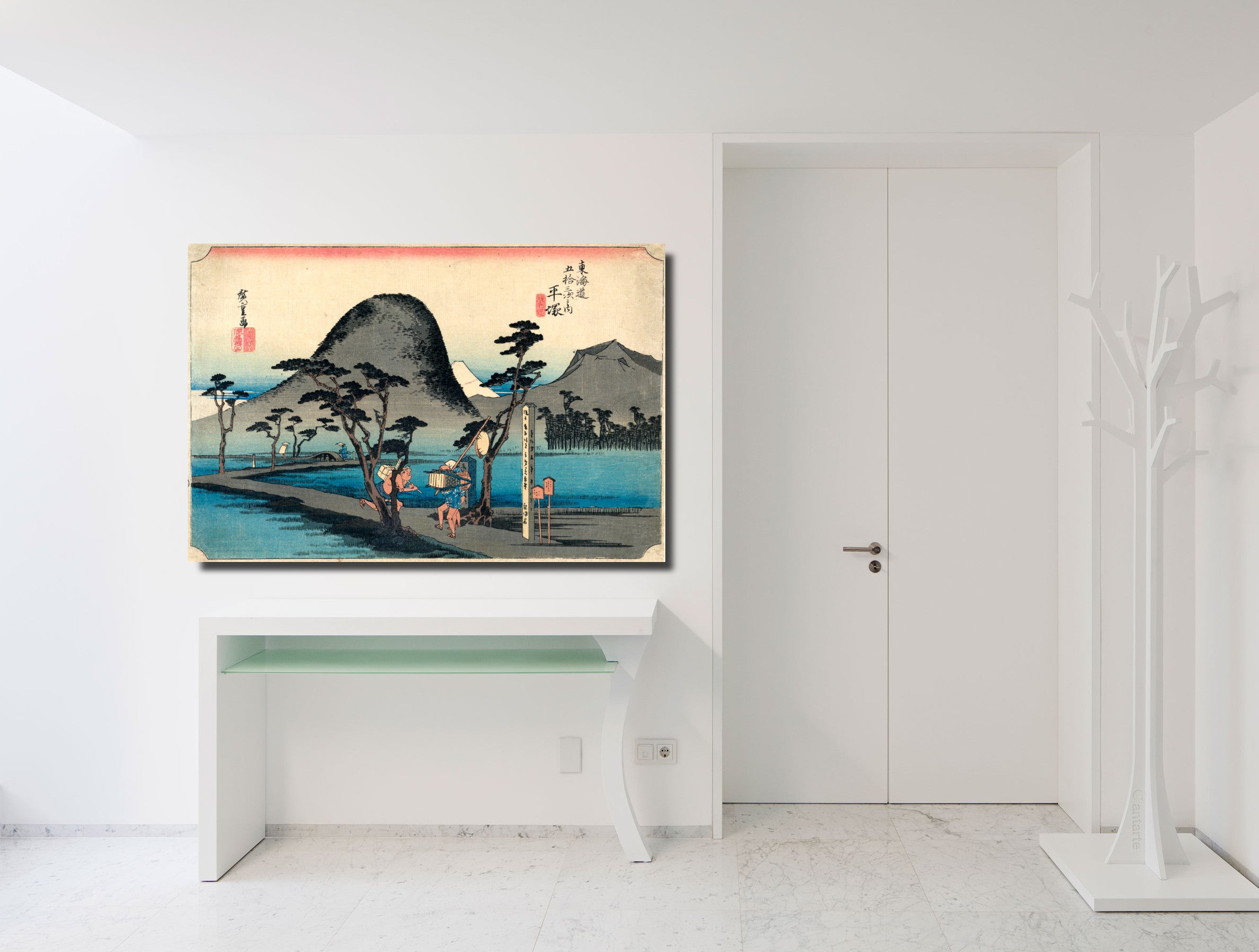 A framed Japanese-style landscape art print hangs on a white wall above a white console table in a bright, modern room. The artwork depicts a scenic coastal landscape with mountains, trees, figures, and blue water, with a gold-toned frame.