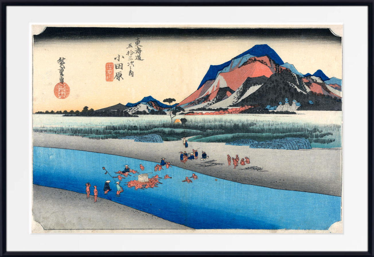A framed Japanese-style landscape print showing a blue river, sandy shore, reeds, and a mountain with red, orange, and blue tones. The artwork includes black calligraphy and red seals, presented with a white mat and black frame.