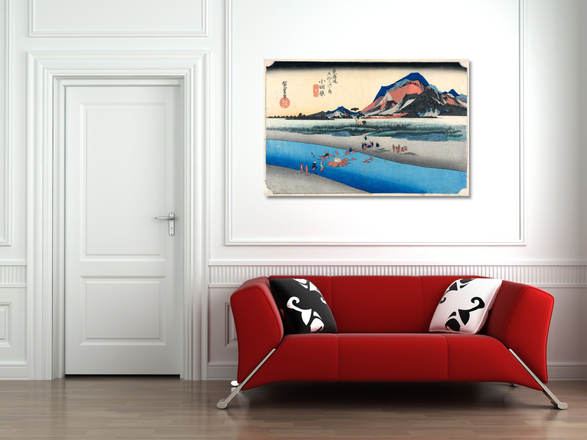 A framed Japanese-style art print depicting a scenic landscape with blue water, mountains, and a red sun, displayed on a white wall above a modern red sofa with black-and-white patterned pillows.
