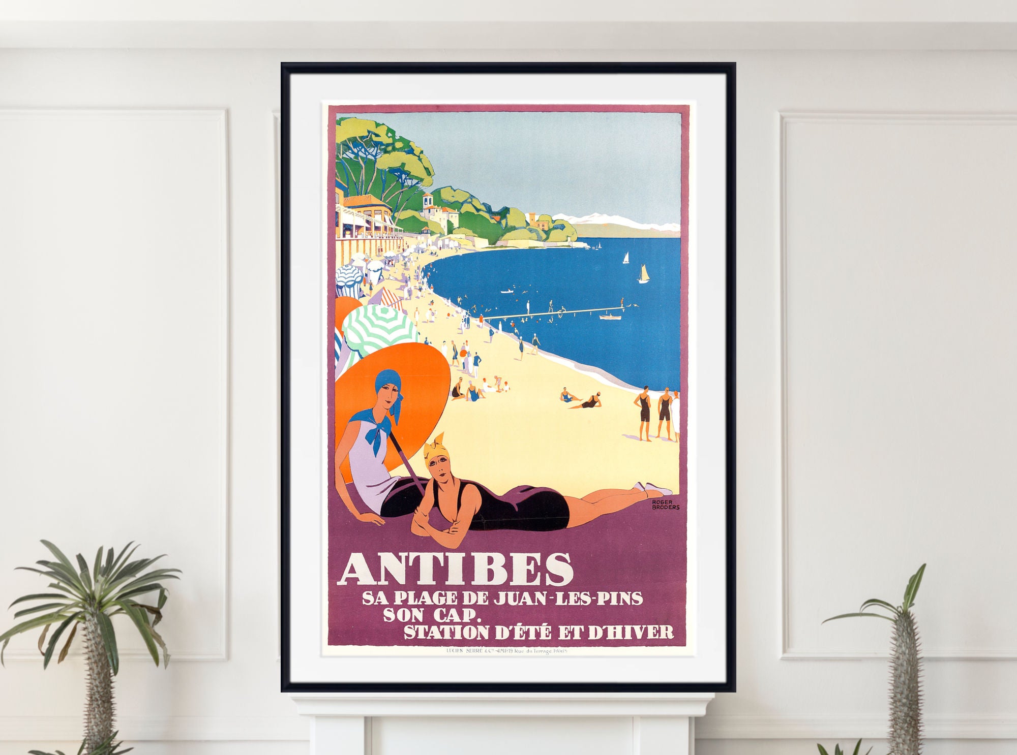 A framed vintage-style travel poster for Antibes beach hangs on a white wall above a mantel, with small potted plants at the sides. The poster shows illustrated beachgoers, a blue sea, sandy shore, and French text reading 'ANTIBES'.