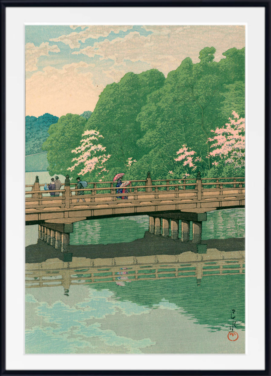 A framed Japanese-style art print depicting a scenic landscape with a wooden bridge over calm water, green trees, pink blossoms, and a pale sky. The print is shown with a white mat and a black frame.