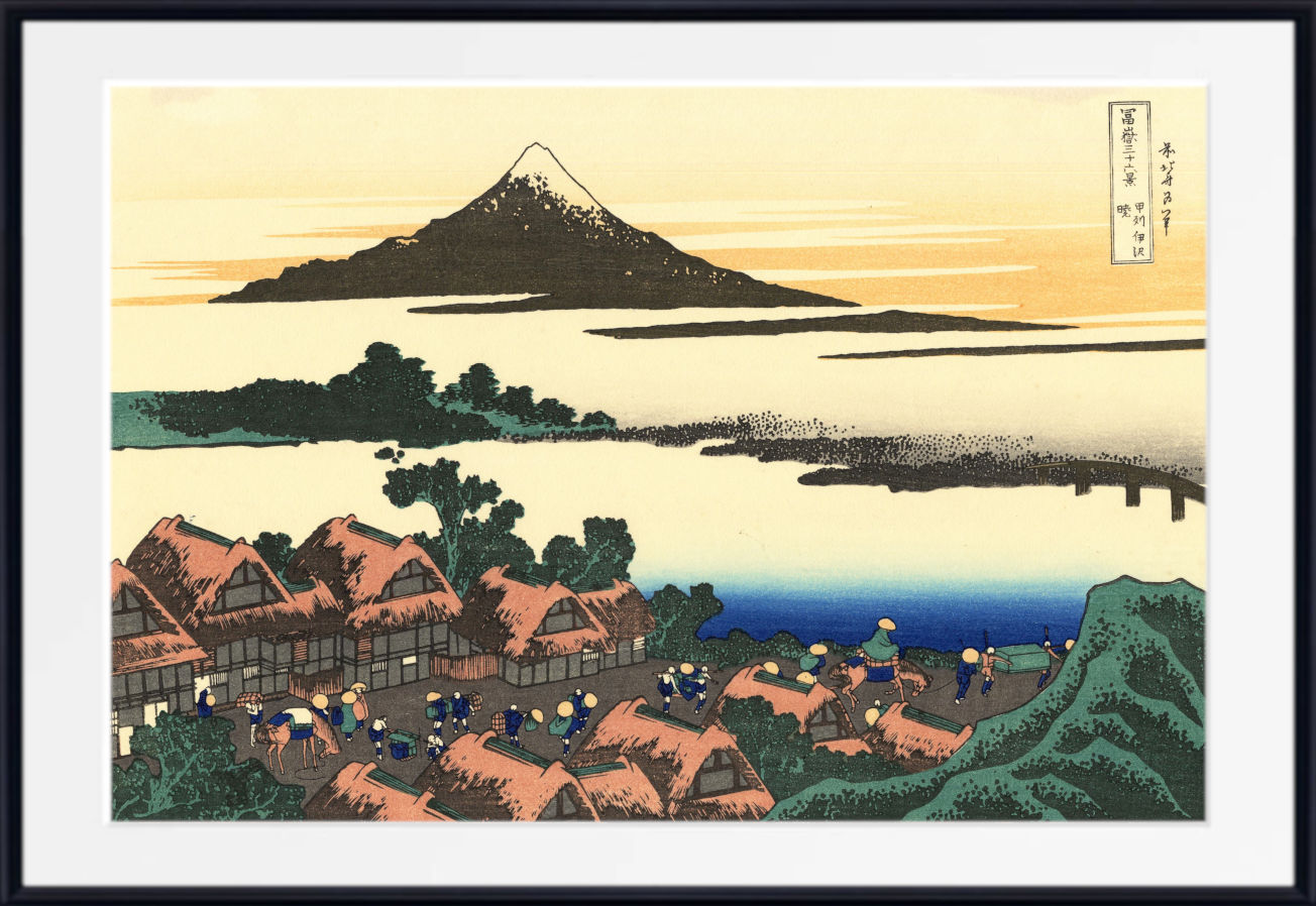 A framed Japanese-style art print depicting a coastal village with thatched-roof houses, a blue sea, and a mountain island in the distance, shown against a white mat and black frame.