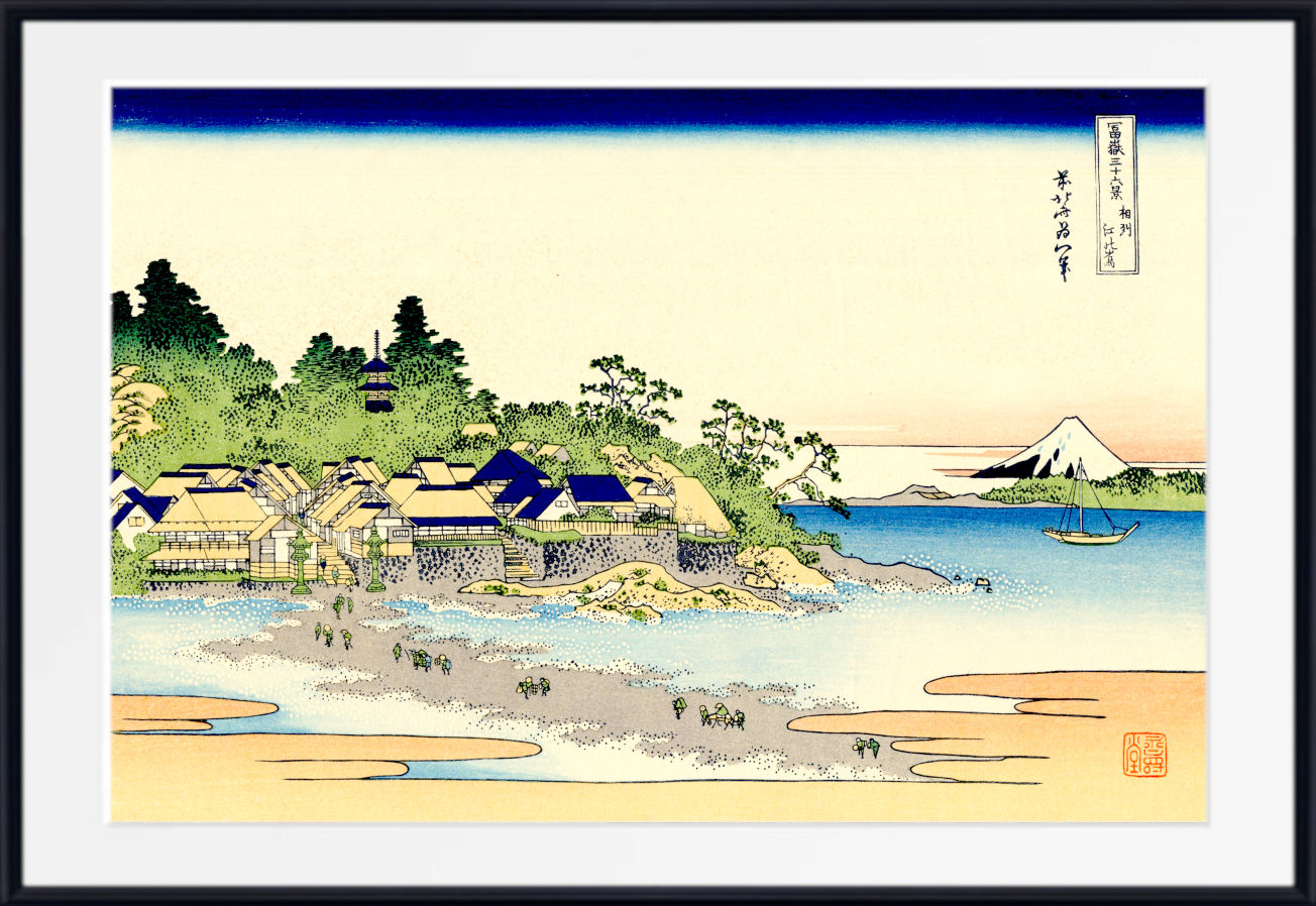 A framed Japanese ukiyo-e woodblock print depicting a coastal village with blue-roofed houses, sandy shore, trees, boats, and a distant mountain, shown with a white mat and black frame.