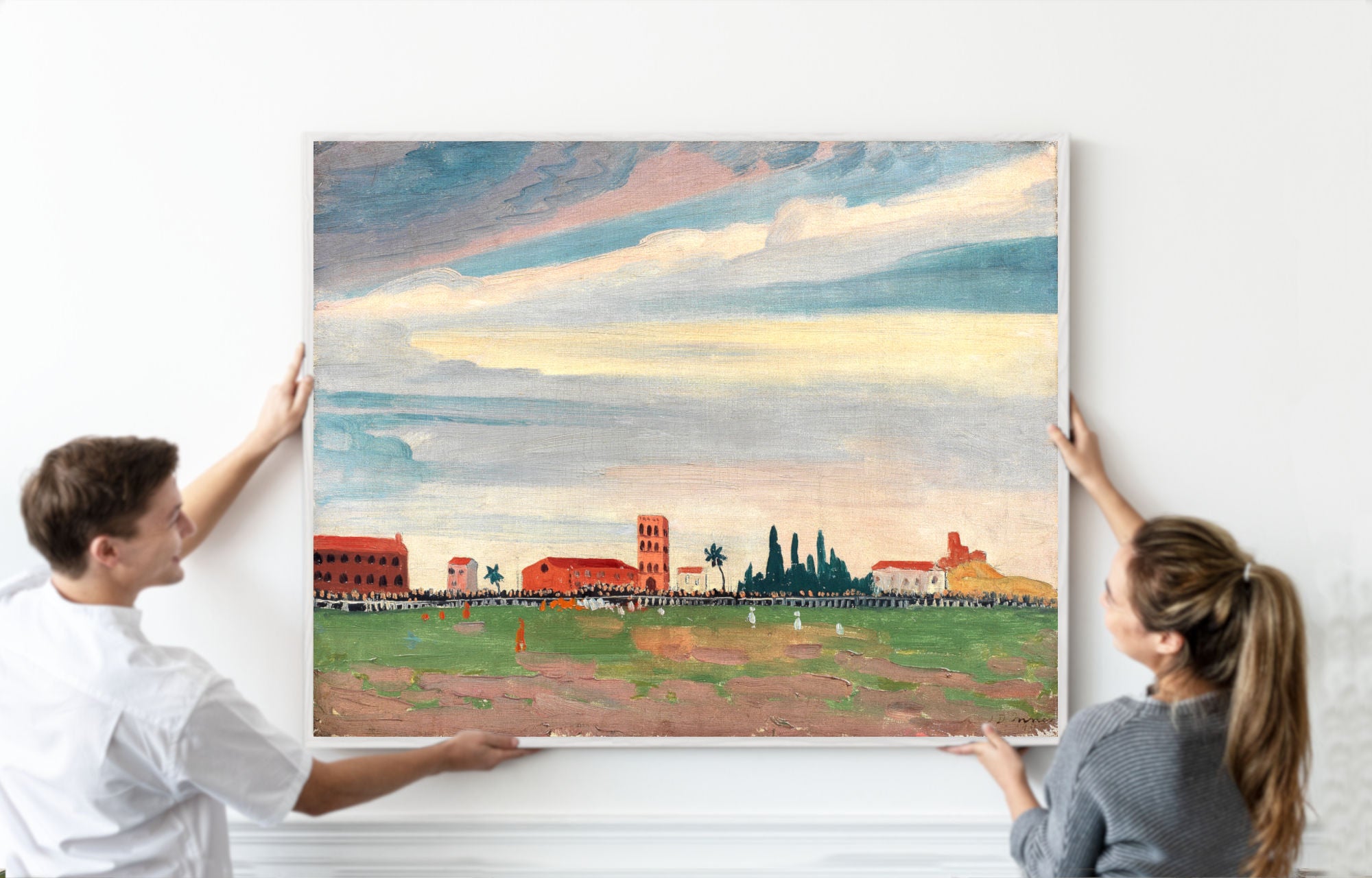 A man and a woman holding up a large framed landscape painting of a stadium under a cloudy sky, set against a clean white wall.