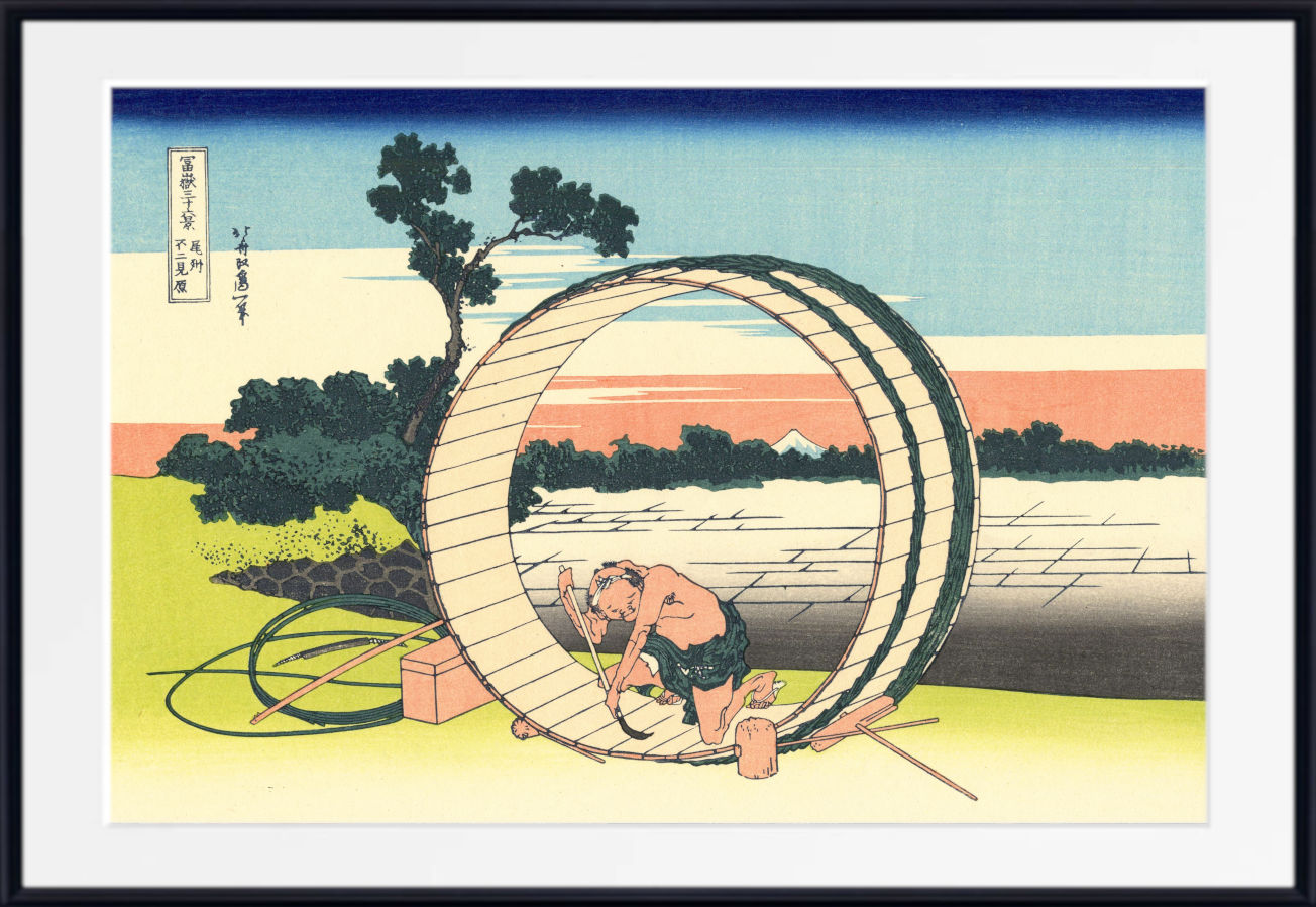 A framed Japanese-style art print showing a man working inside a large circular wooden structure near a lake, with trees, sky, and distant buildings. The print is presented with a white mat and black frame.