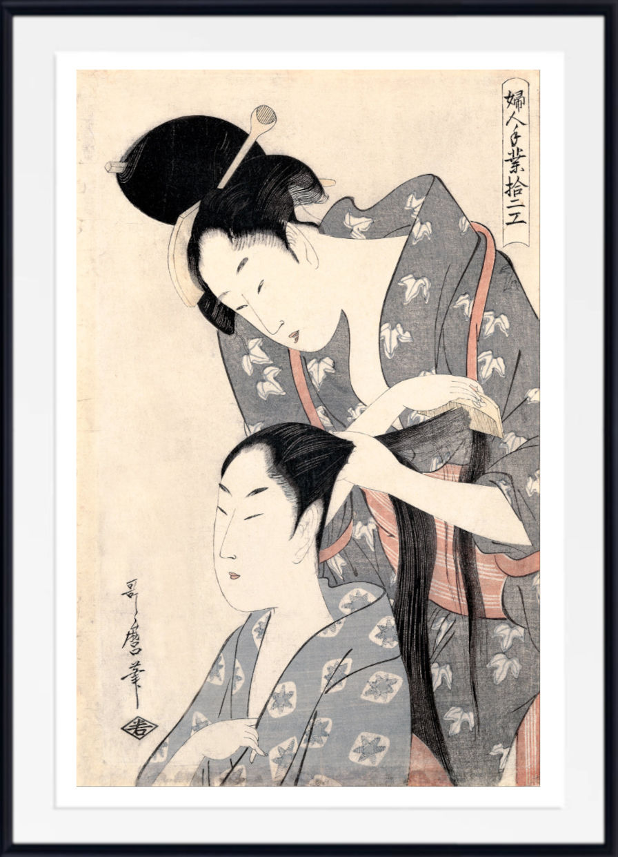 A framed Japanese ukiyo-e woodblock print showing two women in patterned kimonos, with one adjusting the hair of the other. The print is mounted with a white mat and a black frame.
