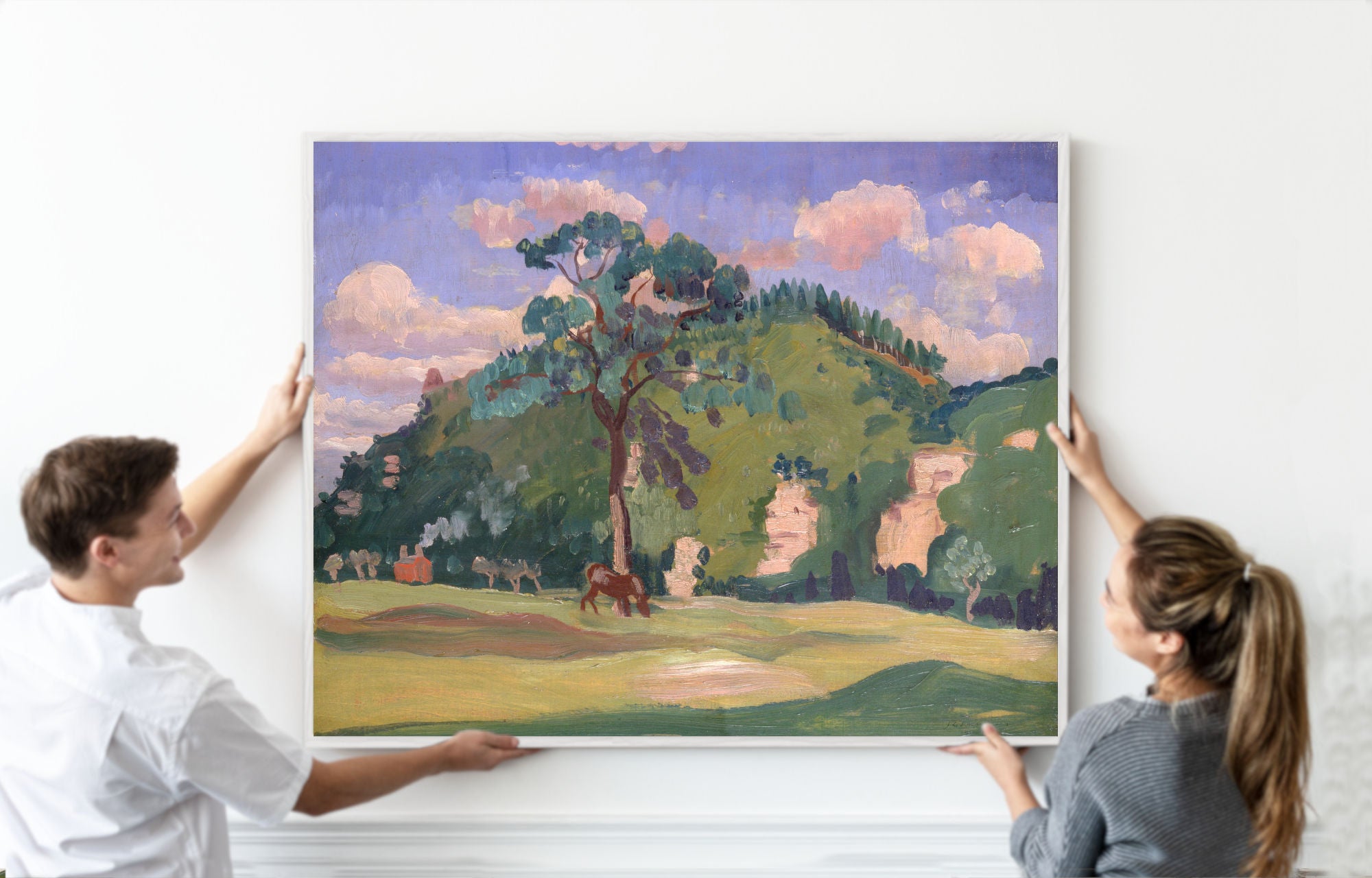 A man and a woman holding a large framed landscape painting of a green hillside with trees, grassy fields, and pinkish clouds under a purple-blue sky, against a white wall.