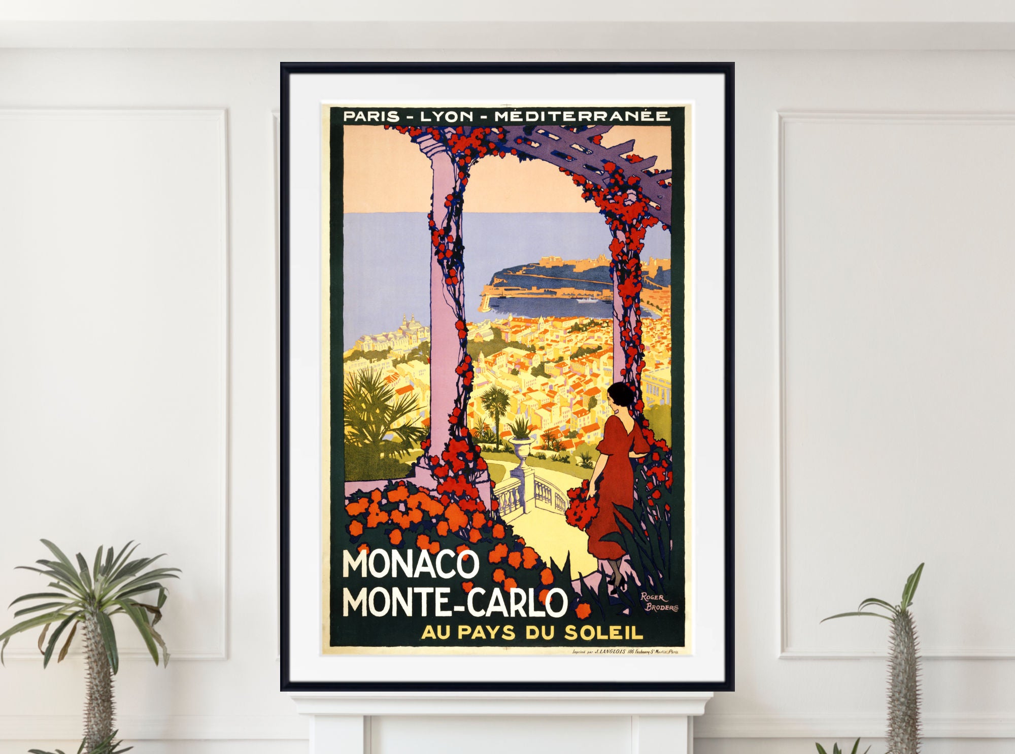 A framed vintage travel poster for Monaco Monte-Carlo hangs on a white wall above a white mantel, flanked by small potted plants. The poster features a woman in a red dress under a floral archway looking out over a coastal landscape, with French text including 'MONACO MONTE-CARLO' and 'AU PAYS DU SOLEIL'.