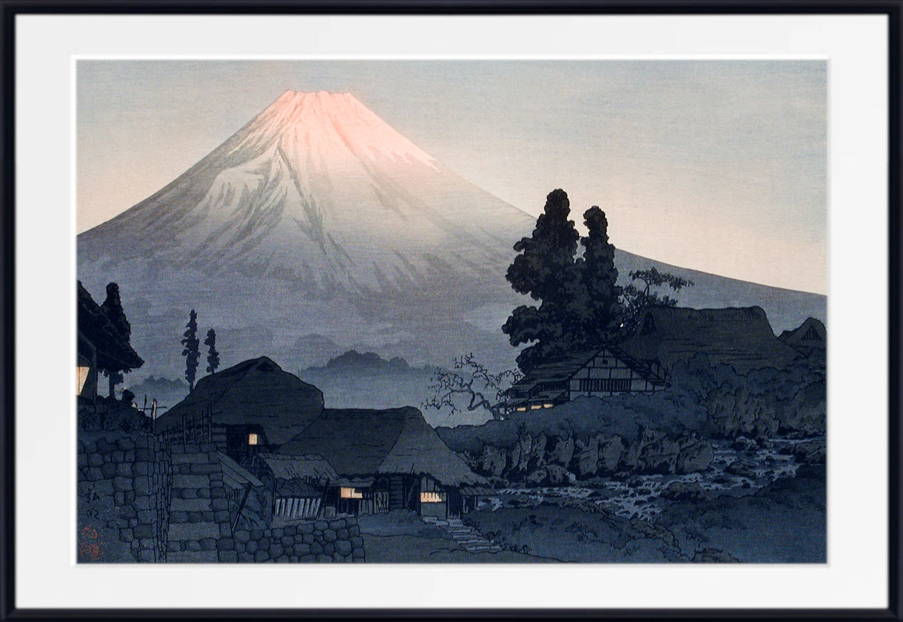 A framed Japanese-style art print depicting Mount Fuji with a pale pink peak, dark trees, stone buildings, and a stream in the foreground, shown with a white mat and black frame.