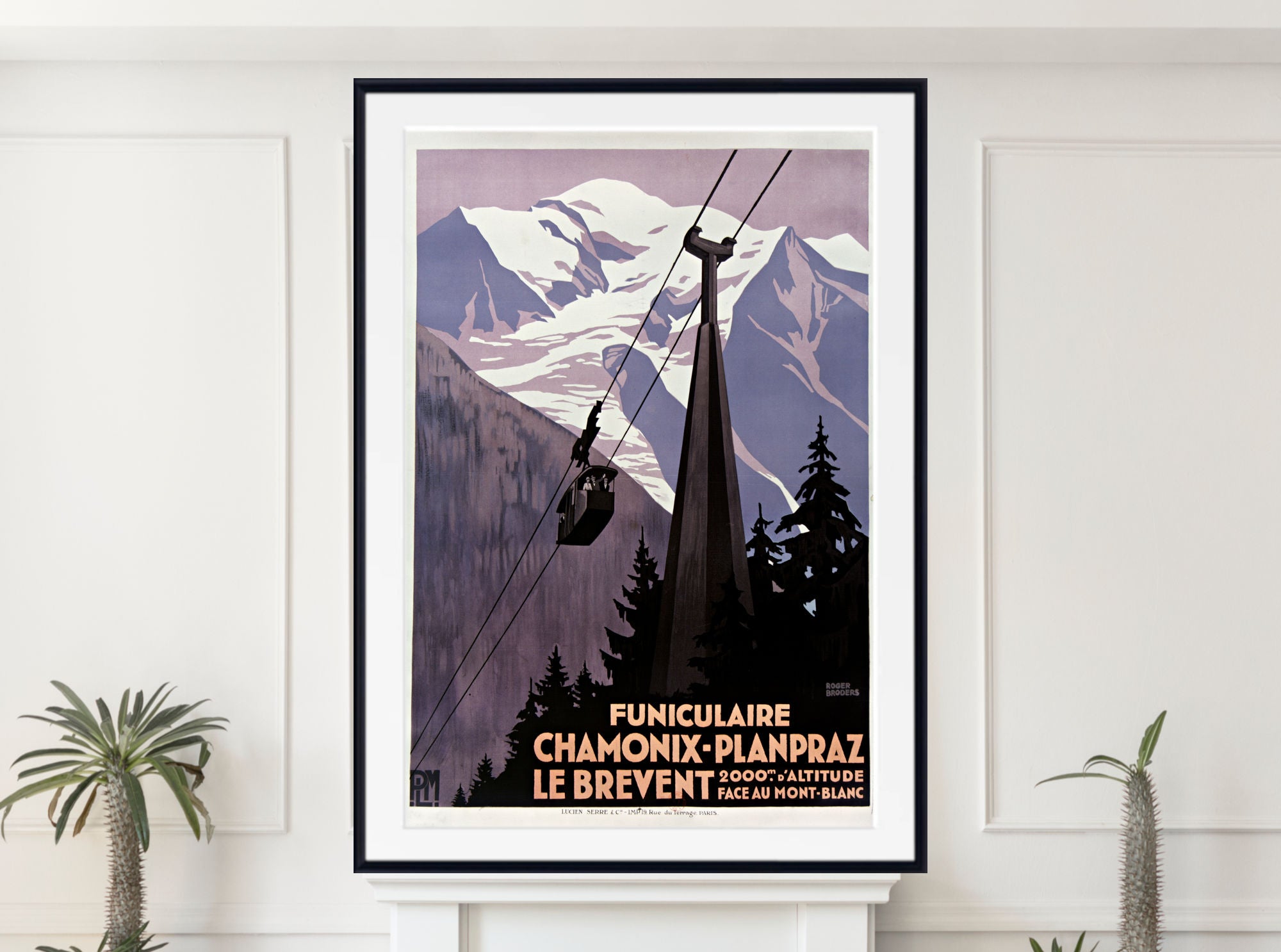 A framed vintage-style mountain cable car poster hangs on a white wall above a mantel, with small potted plants at the sides. The artwork shows snowy mountains, dark evergreen trees, and a cable car silhouette with French text at the bottom.