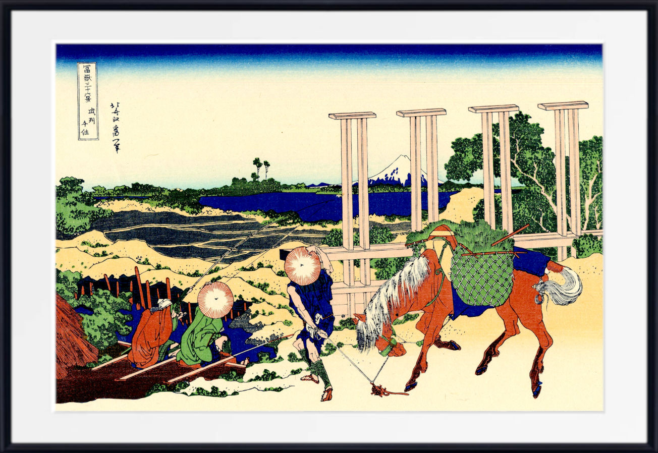 A framed Japanese woodblock-style art print showing figures near a river and large stone pillars, with a blue sky and pale ground. The print is centered within a white mat and a black frame.