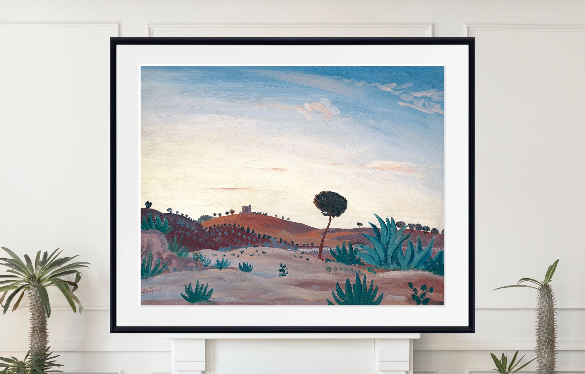 A framed landscape art print featuring a stylized desert scene with a lone tree, cacti, and rolling hills under a blue sky, displayed on a white wall above a mantel with potted plants.