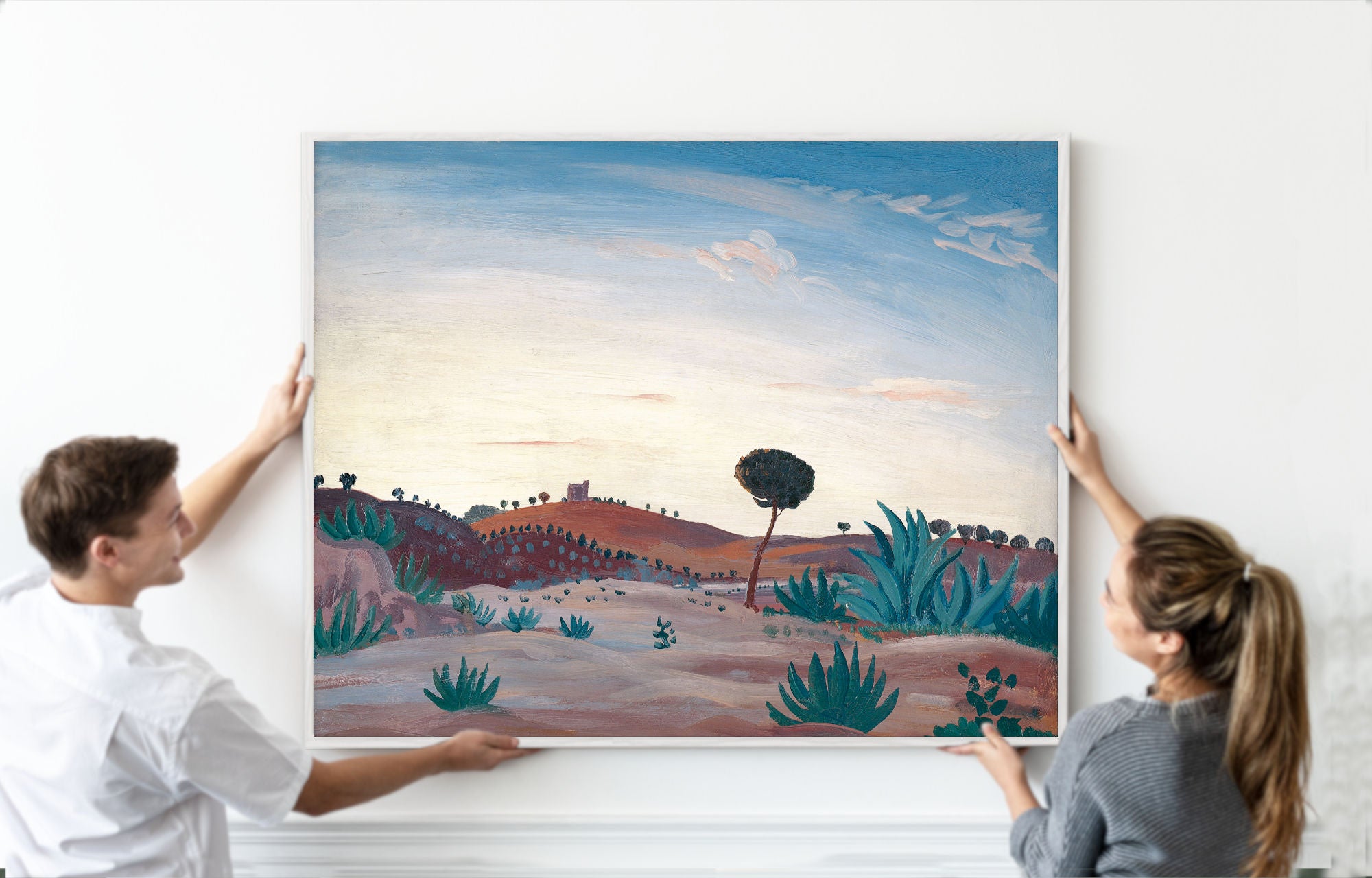 A couple holds up a large framed landscape painting of a desert scene with green cacti and a blue sky, set against a clean white wall.