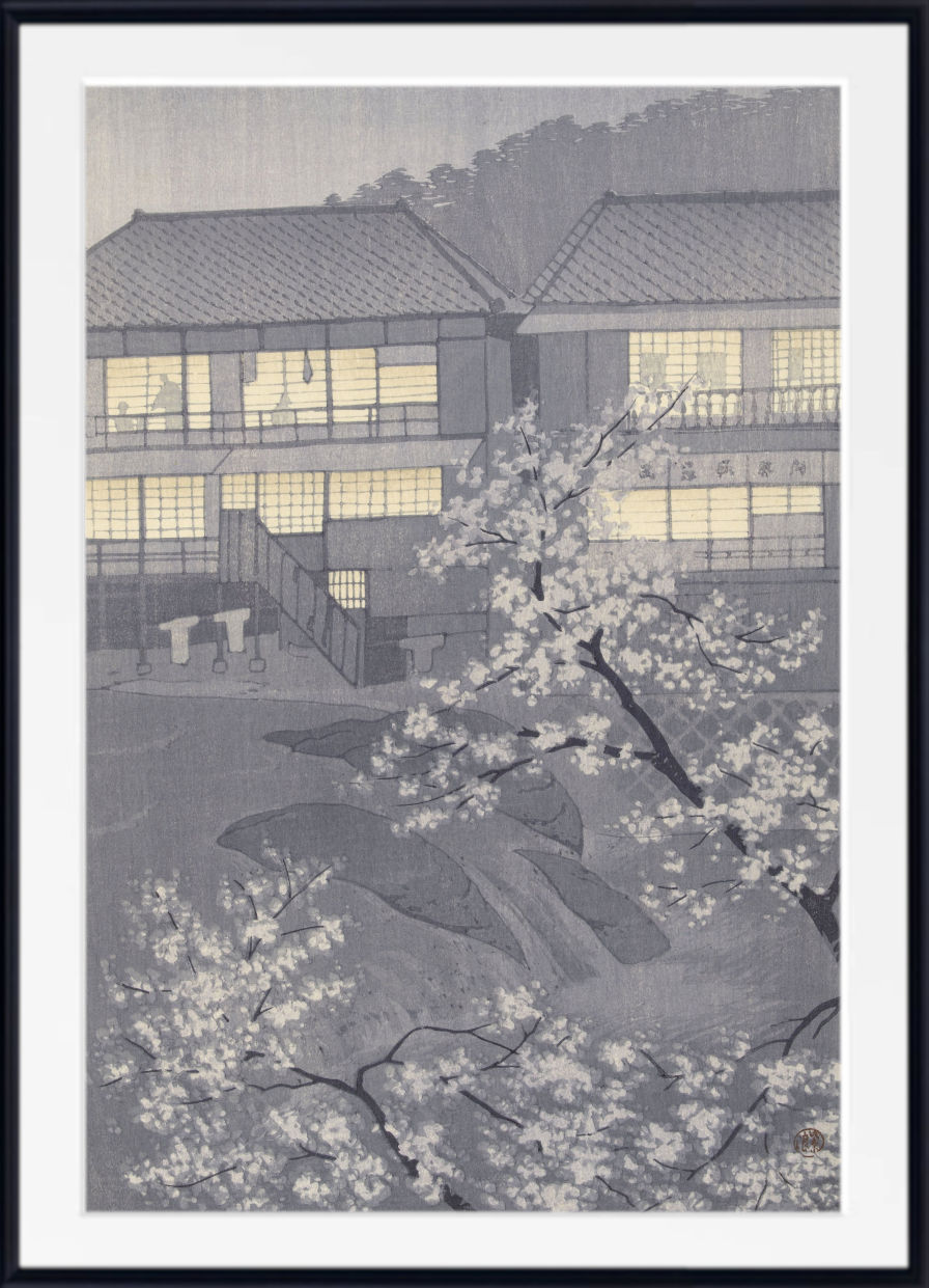 A framed Japanese-style art print depicting a traditional building with glowing windows and white flowering cherry blossoms in the foreground, shown against a dark background.