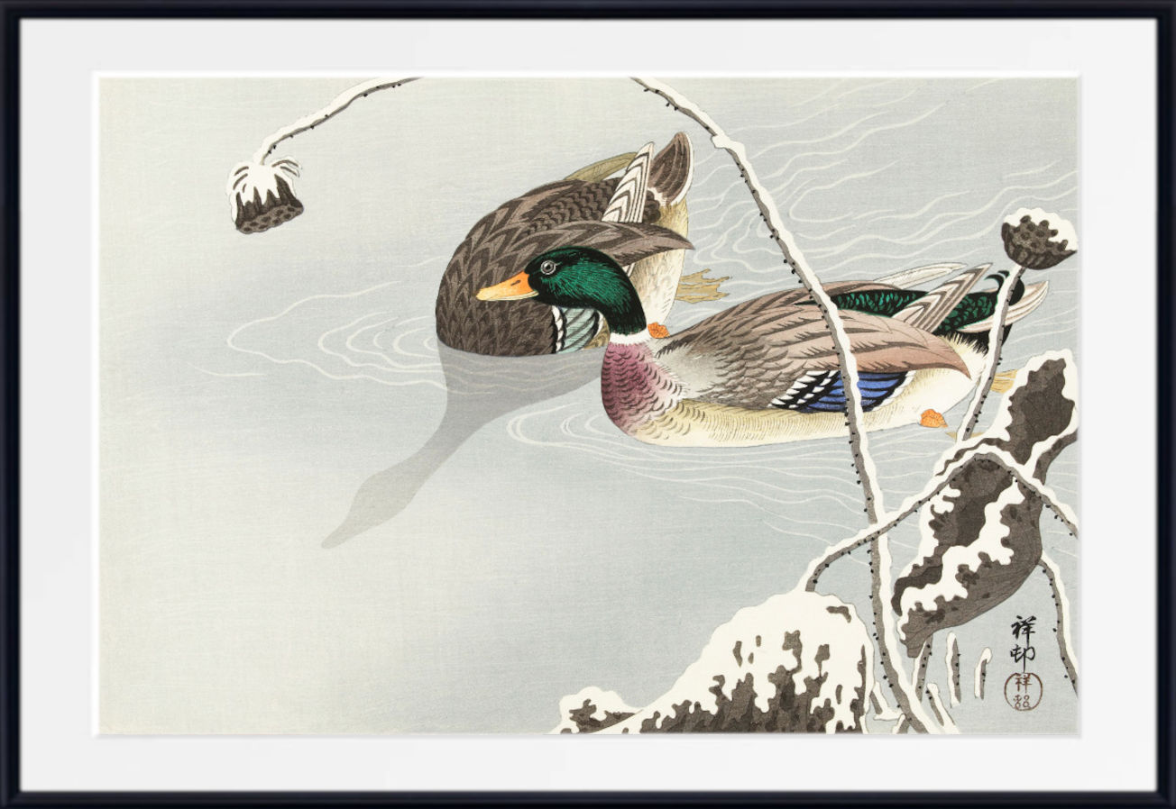 A framed Japanese-style art print depicting two mallards swimming in a pale winter pond with snow-covered branches and dark rocks, shown against a white background.