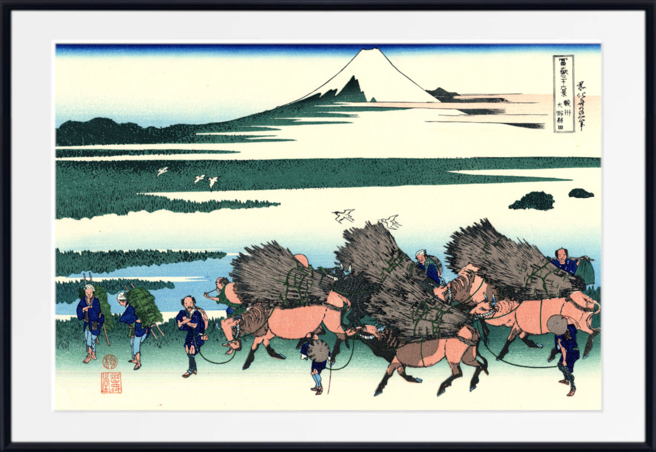 A framed Japanese woodblock-style art print showing a snowy mountain, water, and figures carrying large bundles of firewood on horseback. The print is mounted with a white mat and a black frame.
