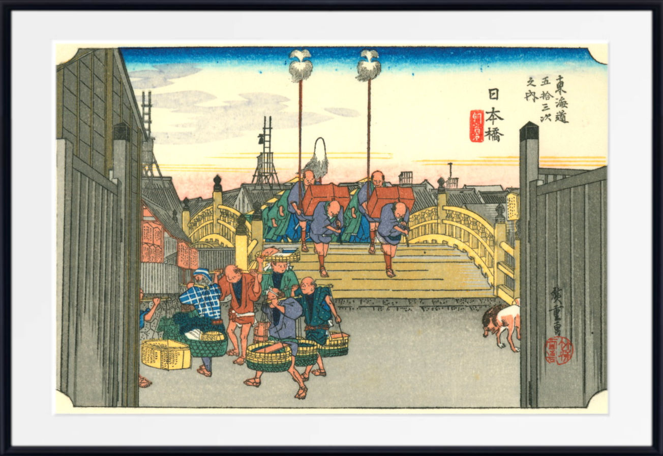 A framed Japanese ukiyo-e woodblock print depicting a street scene with people carrying baskets and goods near a bridge, shown against a white mat and black frame.