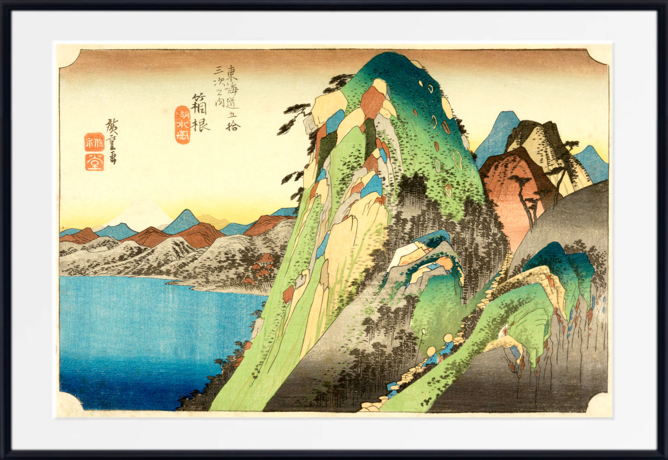A framed Japanese-style landscape print showing a steep green and blue mountain beside a blue body of water, with a cream sky and red seal markings, presented in a black frame with white matting.