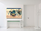 A framed Japanese ukiyo-e woodblock print hangs on a white wall above a white console table in a bright, modern room. The artwork depicts figures in blue and green garments walking through a landscape with trees and mountains.