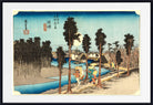 A framed Japanese ukiyo-e woodblock print showing a river scene with tall trees, small boats, houses, and a group of people walking along a path under a pale sky. The print is mounted with a white mat and a black frame.