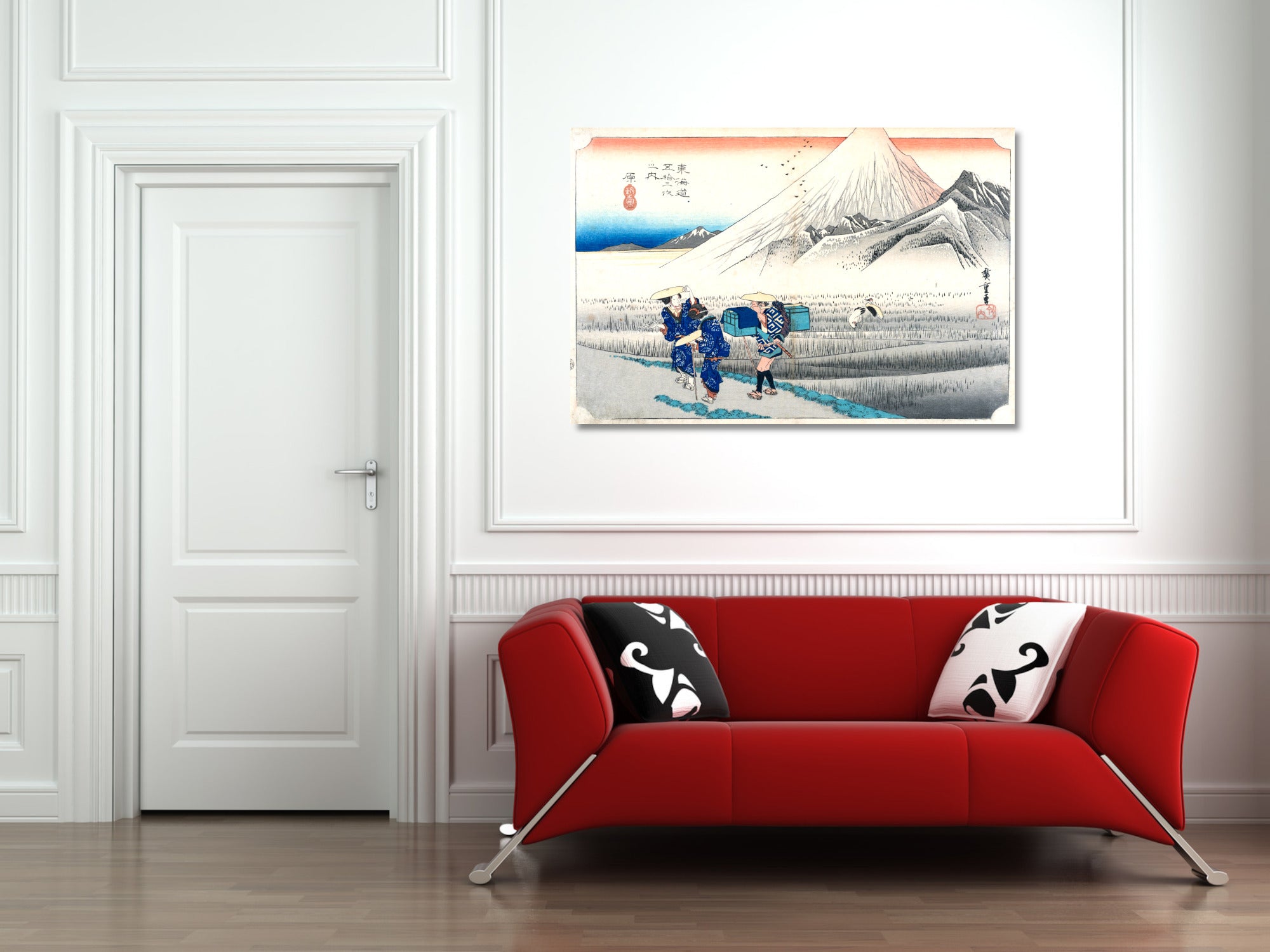 A framed Japanese ukiyo-e style art print depicting Mount Fuji and figures in a landscape, displayed on a white wall above a modern red sofa with black and white patterned pillows.