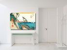A framed Japanese ukiyo-e style landscape art print hangs on a white wall above a white console table in a bright, modern room. The print depicts a scenic coastal landscape with mountains, trees, and blue water, with a prominent yellow band at the top.