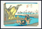 A framed Japanese ukiyo-e woodblock print depicting a coastal scene with figures in a river, a large yellow rock formation, trees, and distant buildings, shown against a white mat and black frame.
