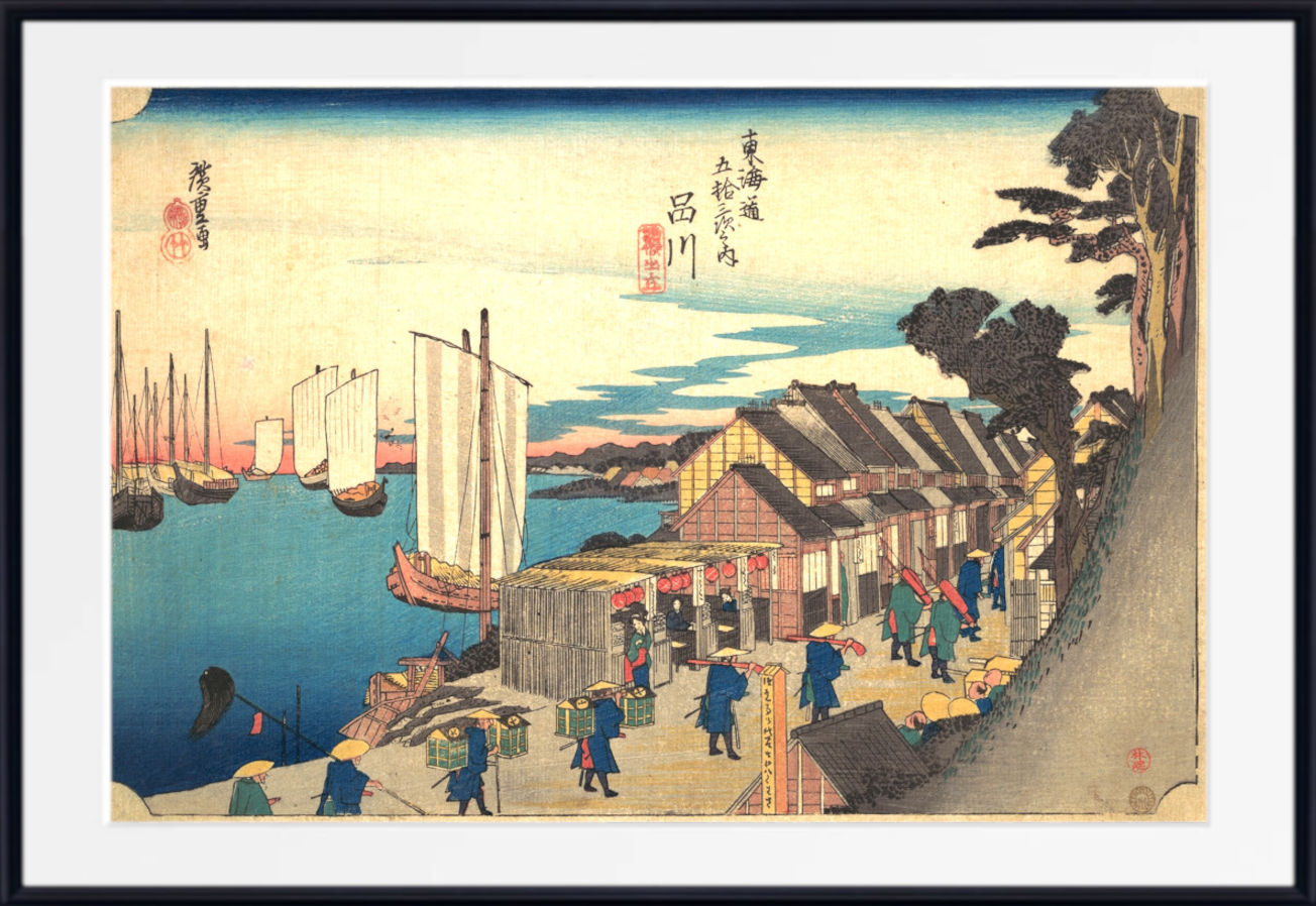 A framed Japanese ukiyo-e woodblock print depicting a harbor scene with boats, buildings, figures, and trees, shown against a white mat and black frame.